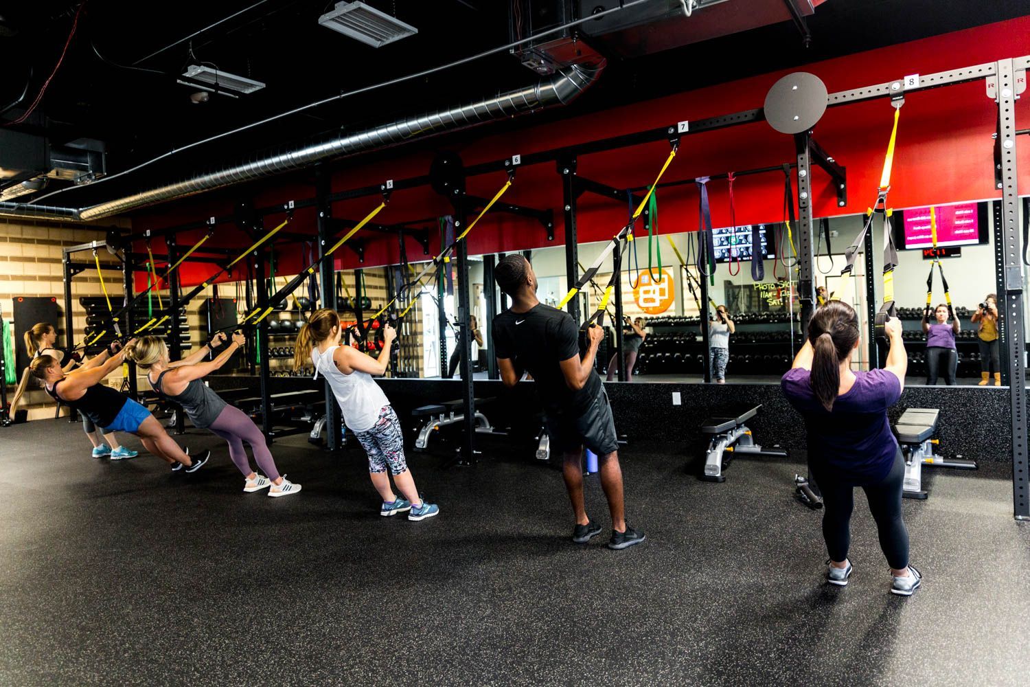People exercising with suspension trainers in a gym. Dark floor, red wall, black equipment, and a mirror.