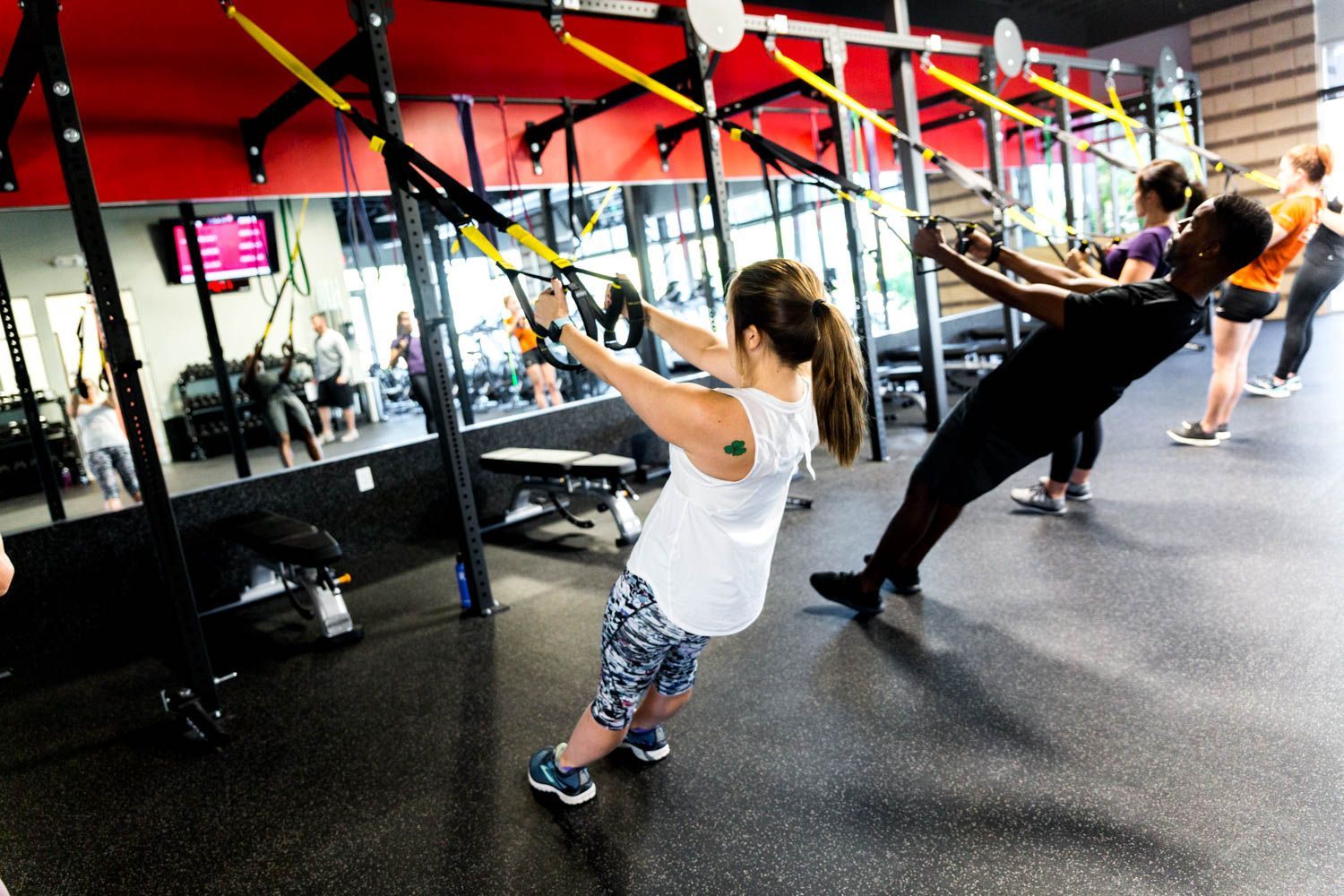 People exercising with TRX straps in a gym, red and black accents, mirrored wall.