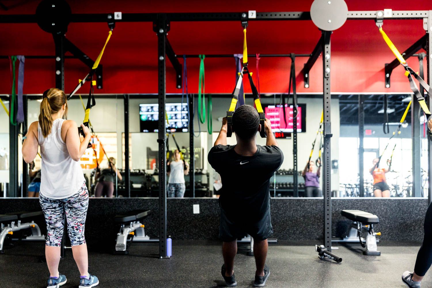 People exercising with suspension straps in a gym, with red accents and a mirrored wall.