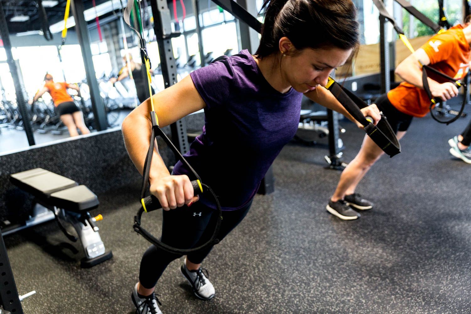 Woman in purple shirt doing a suspension push-up exercise in a gym, with others using the equipment.