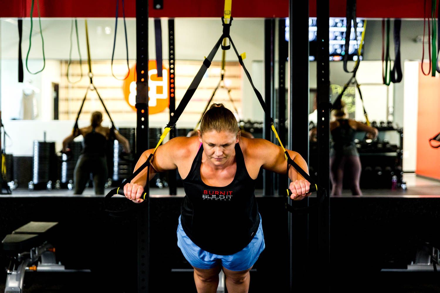 Woman doing a TRX chest press in a gym, pulling on yellow straps, focused expression.