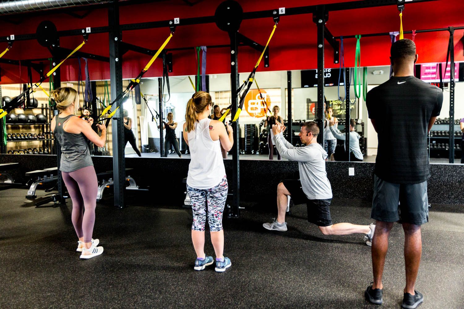 People working out in a gym, using suspension trainers and mirrors. Red and black accents, bright lighting.