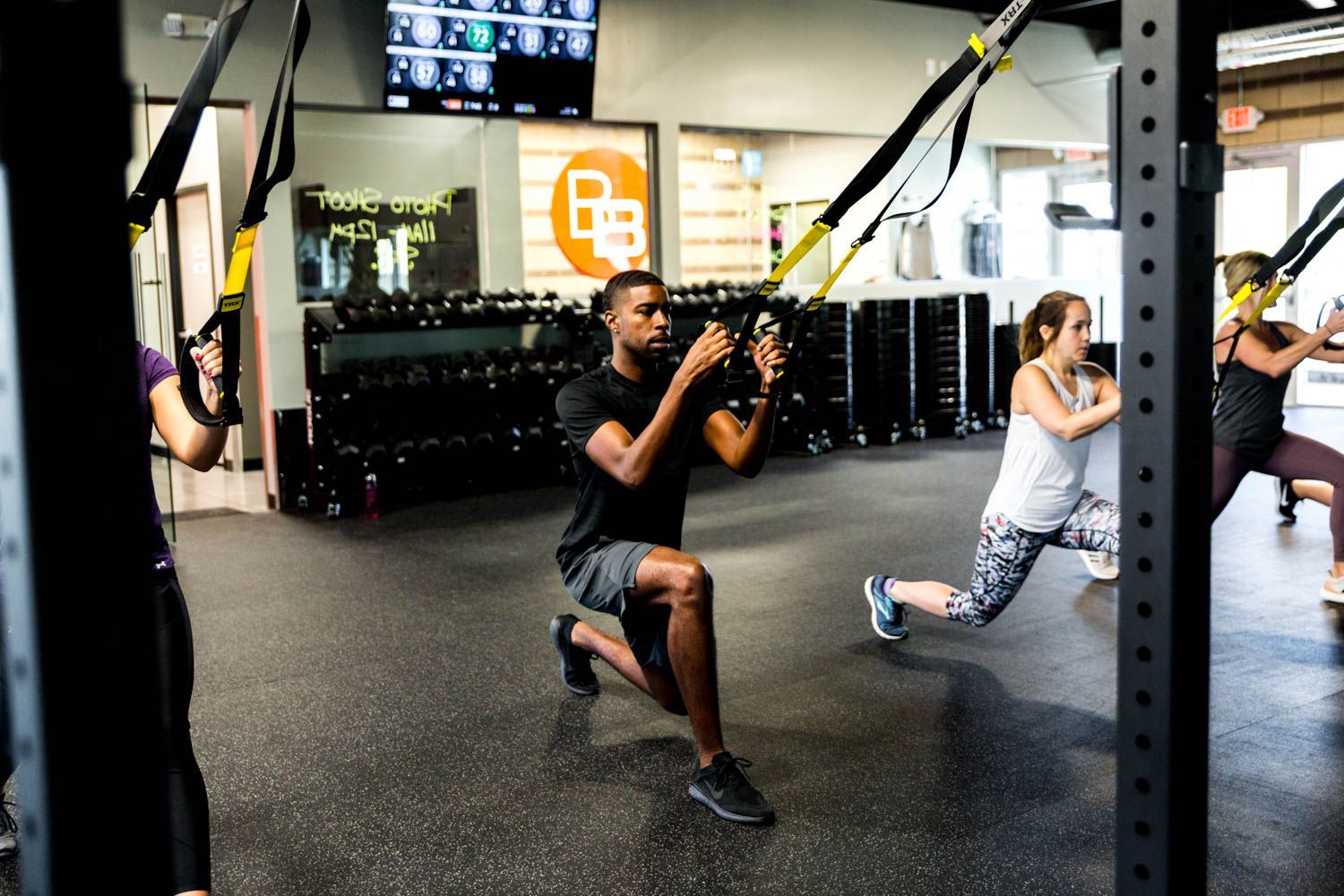 People exercising with suspension trainers in a gym. Man in black shirt doing a lunge.