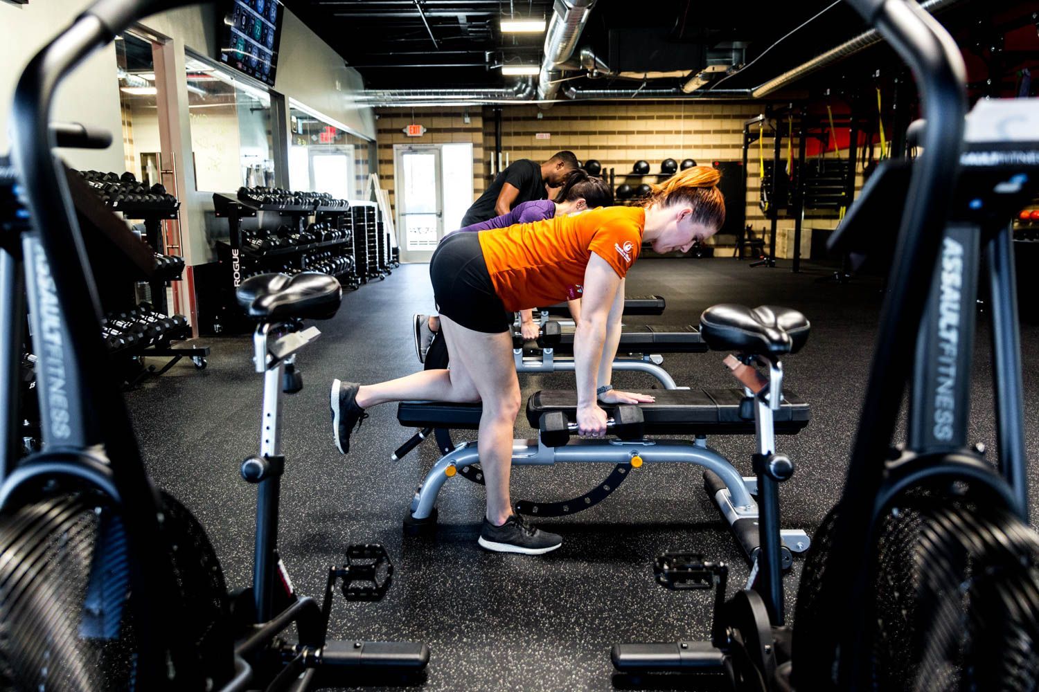 Woman in orange shirt doing a weight exercise in a gym, with exercise bikes in the foreground.