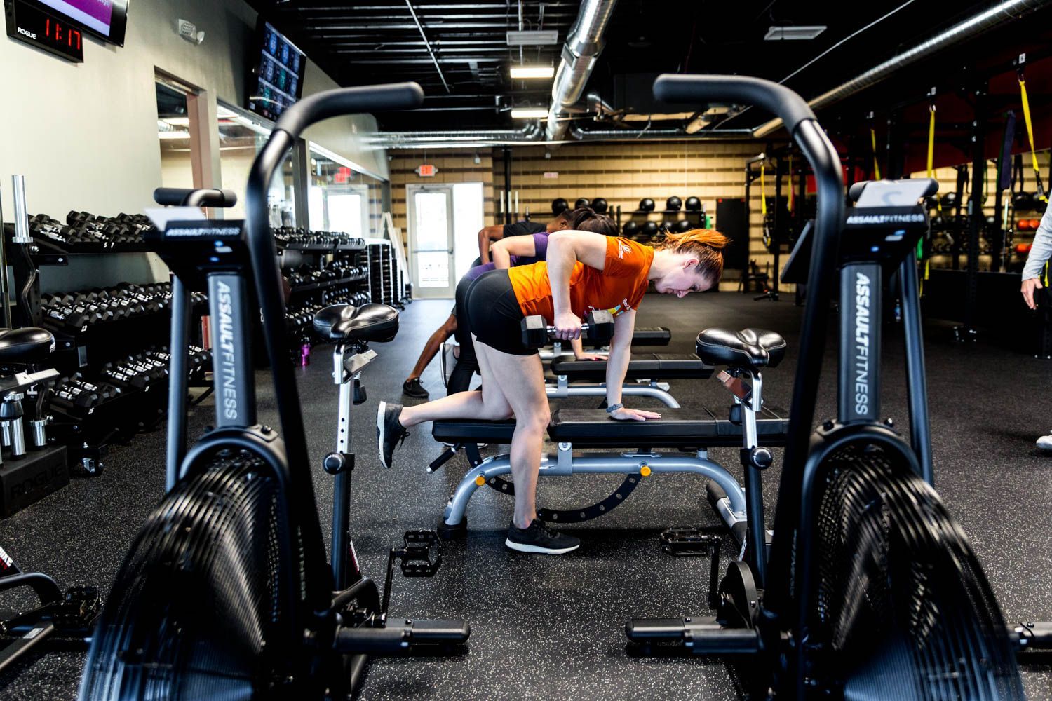 Woman in orange shirt doing a dumbbell row on a bench in a gym, with exercise bikes in foreground.