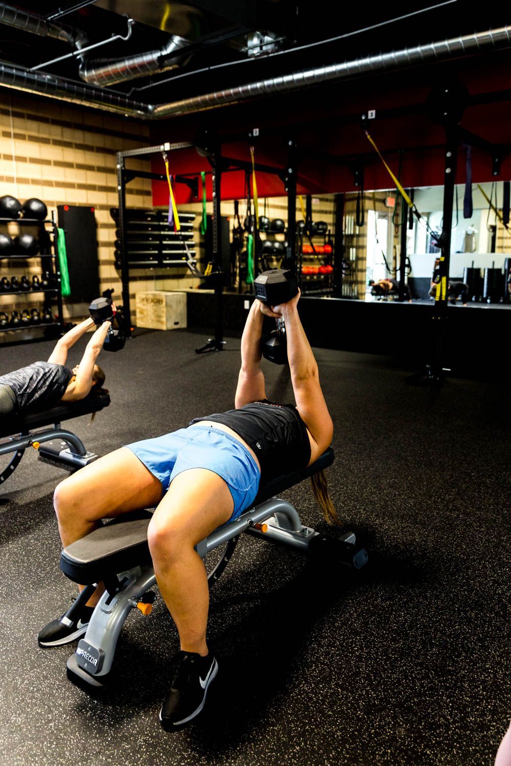 Person on bench lifting dumbbells in gym. Black shirt, blue shorts, dark floor, equipment visible.