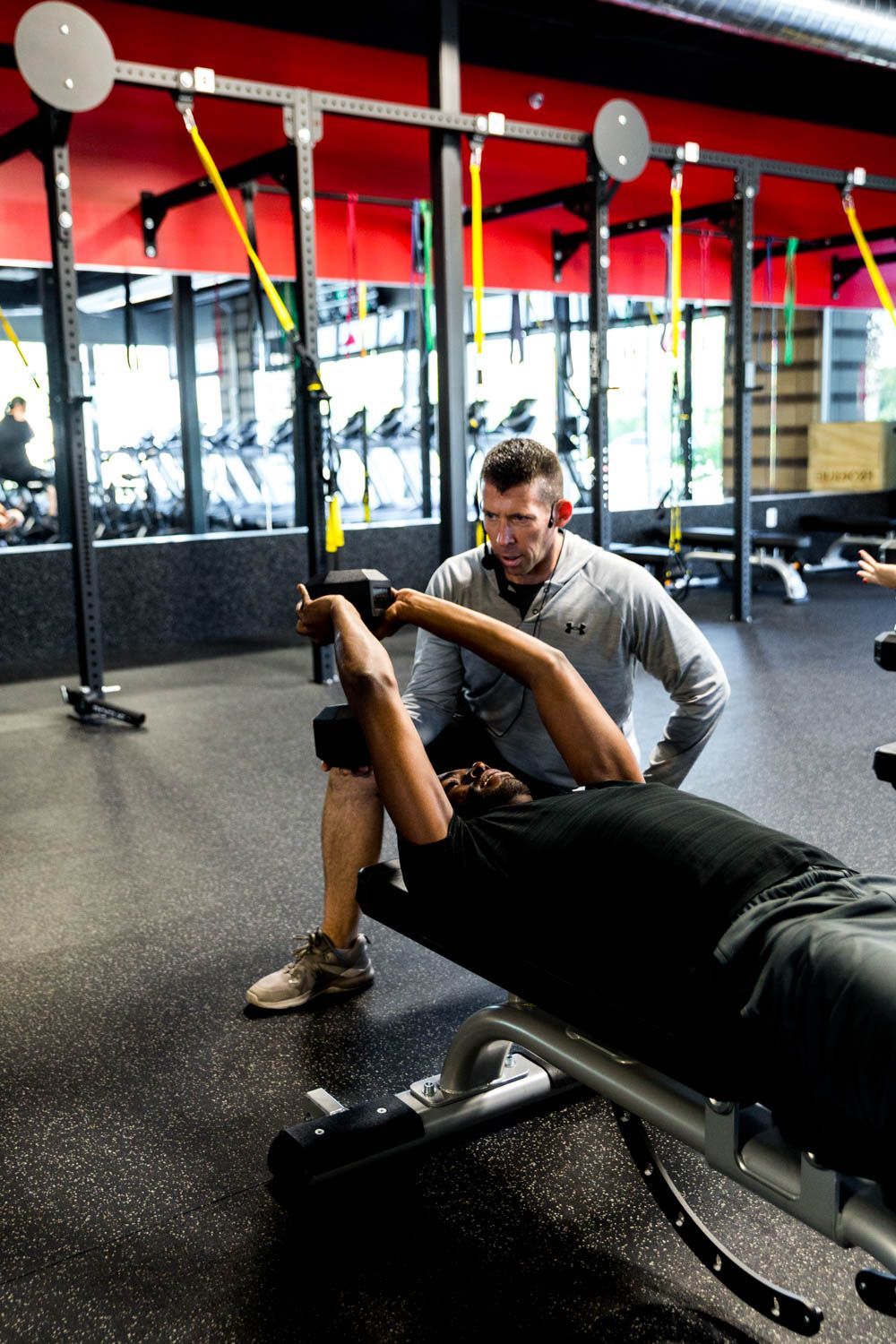 Man doing dumbbell exercises with assistance from a trainer in a gym.