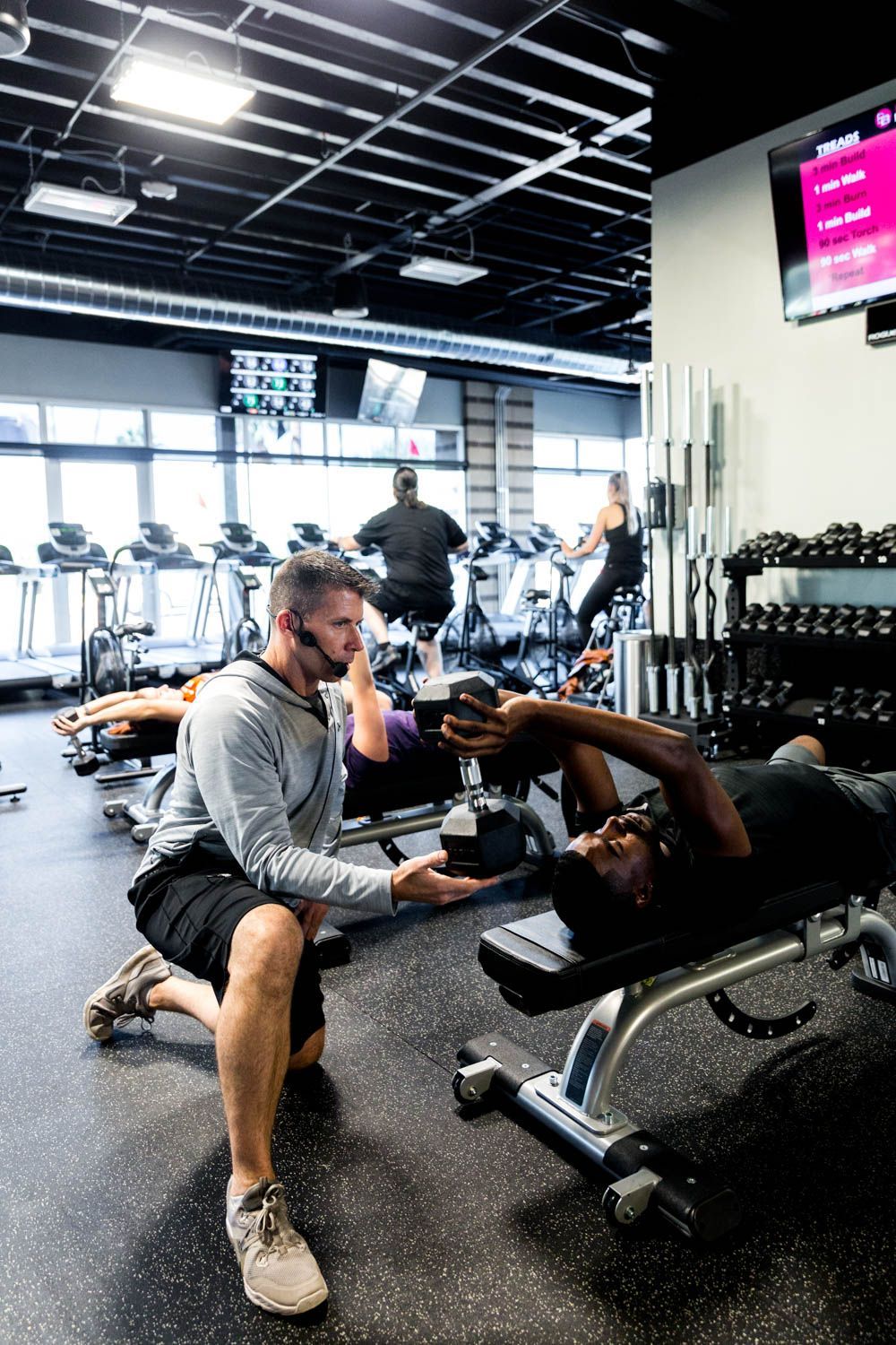 Man kneeling, assisting person using dumbbell on bench in a gym. Other people exercising on treadmills in the background.