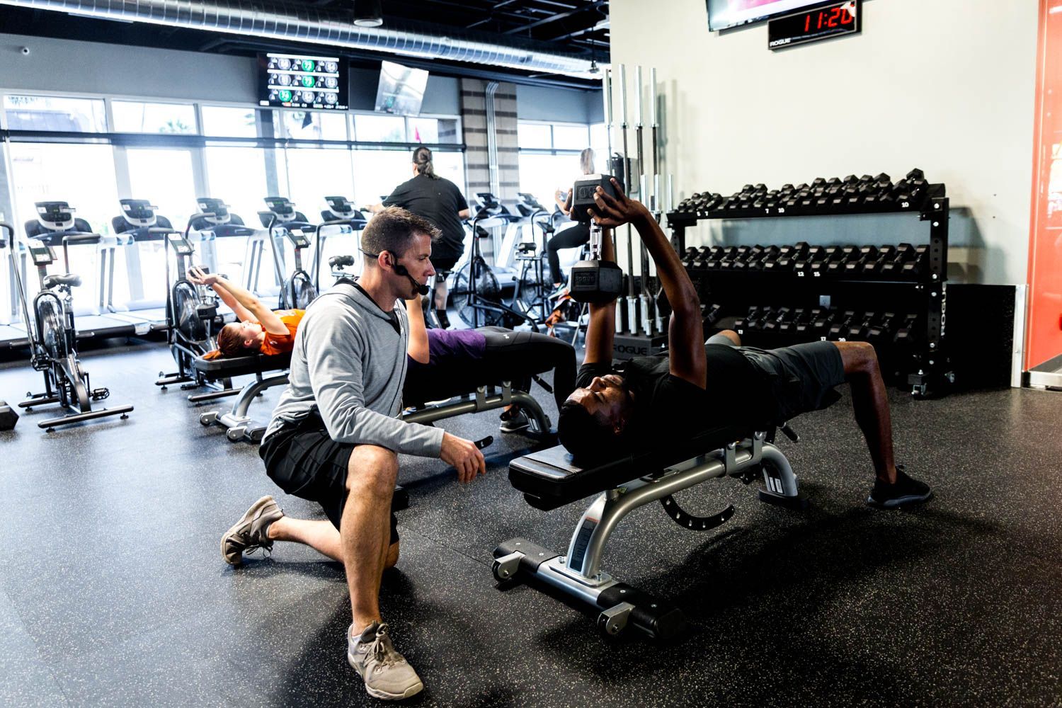 Gym: Man on bench lifting dumbbell, trainer assisting. Others exercise in background.