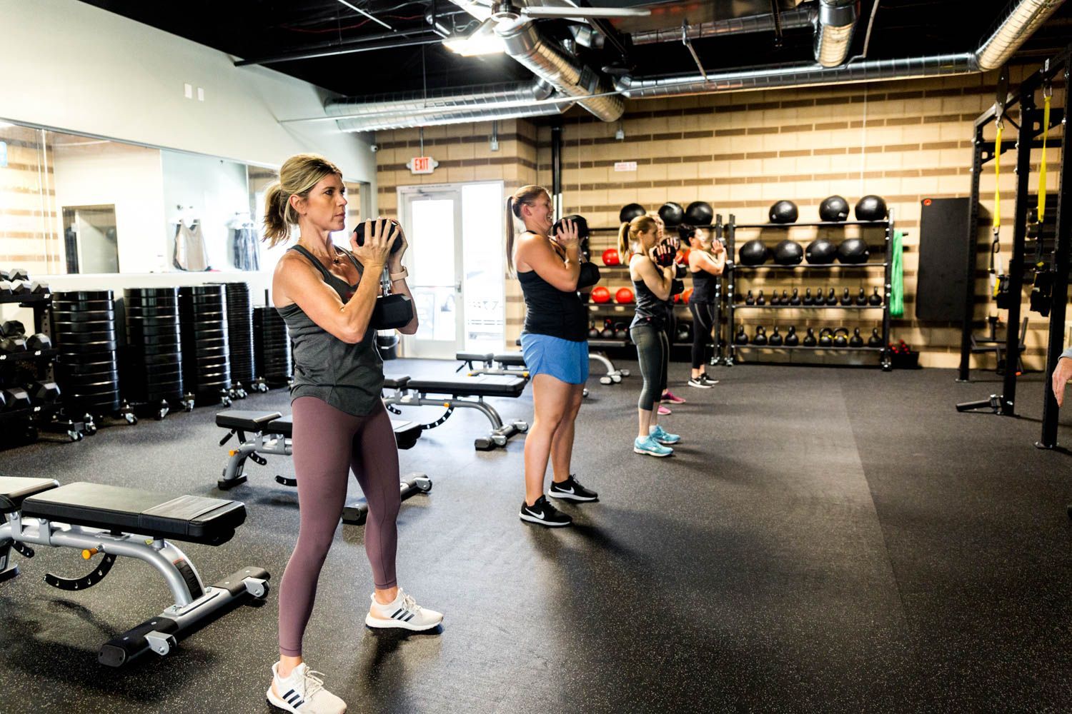 People exercising with kettlebells in a gym. Interior shot with weights, benches, and a black ceiling.