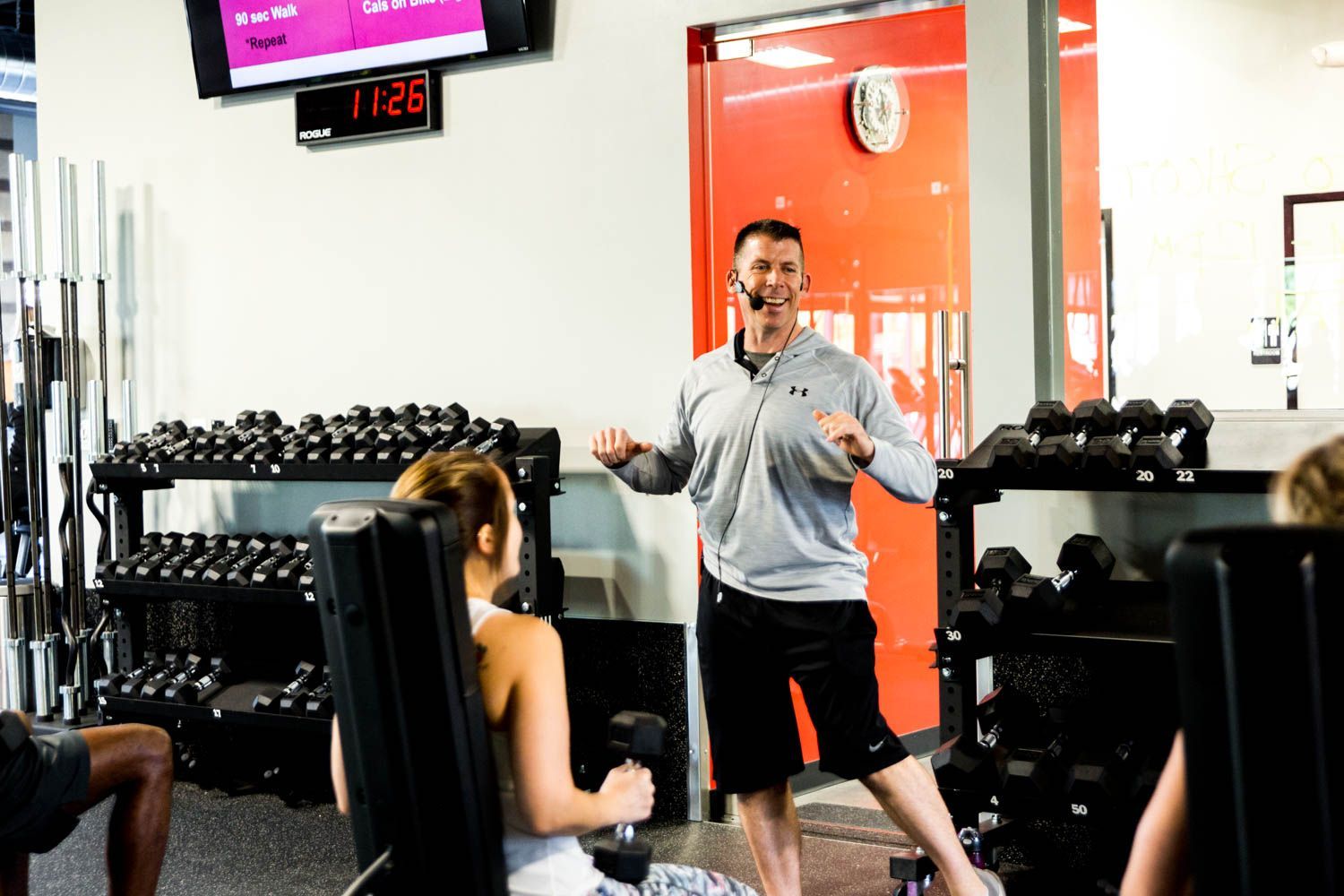 A fitness instructor leads a workout in a gym, with weights and a timer visible.