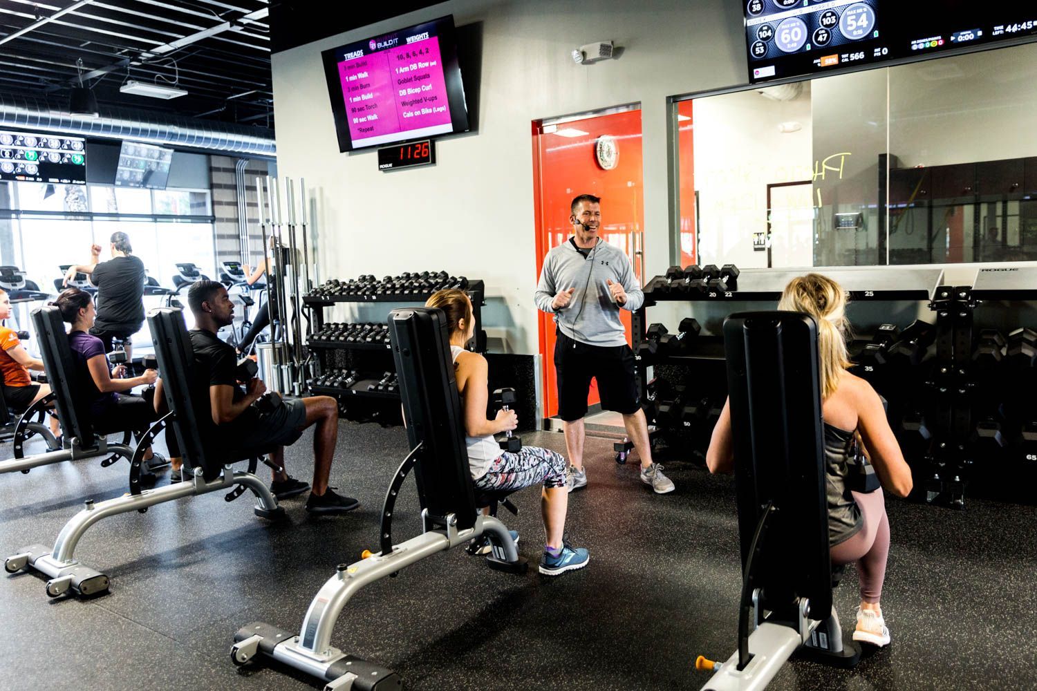 People exercising with weights in a gym, led by a trainer.