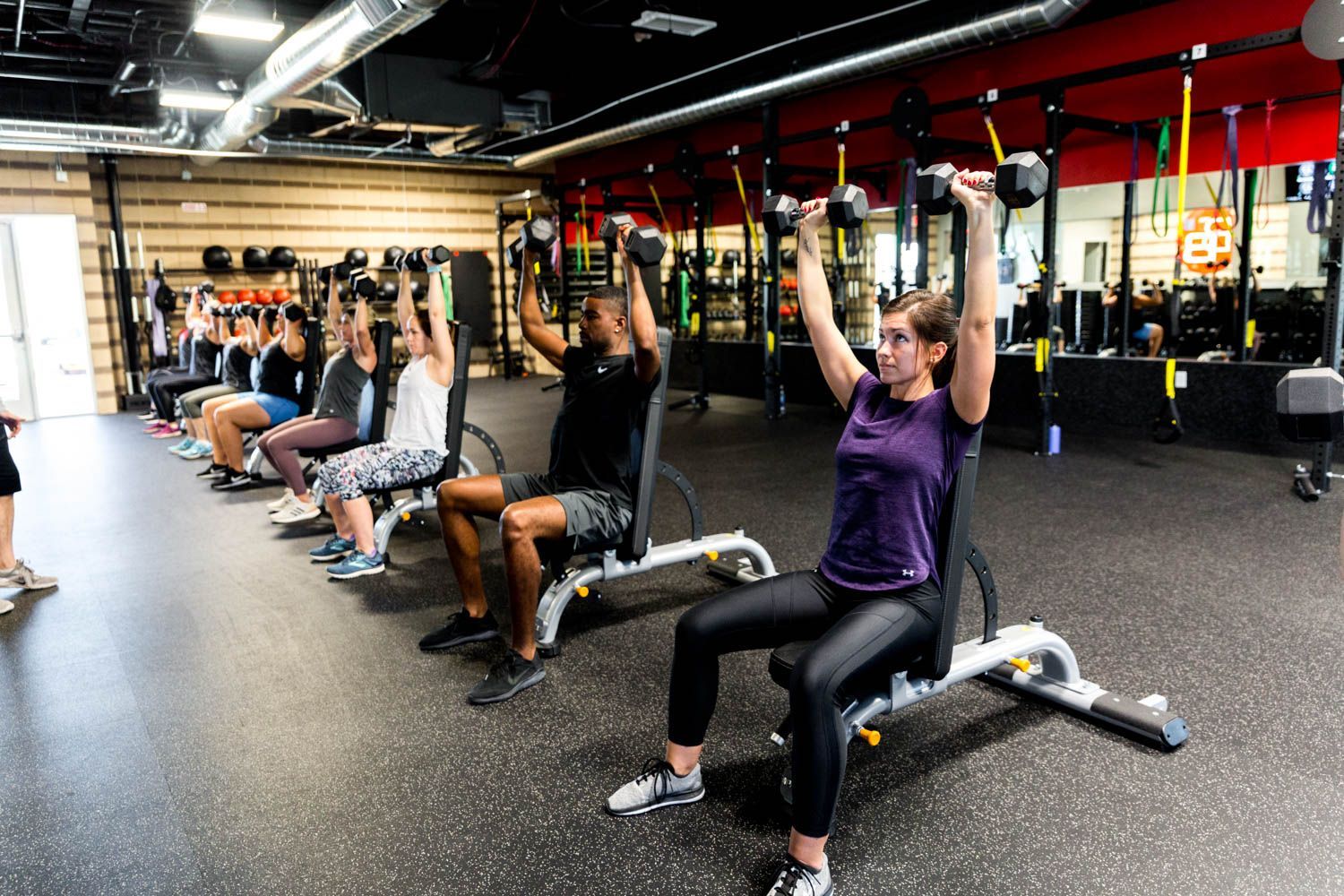 People seated on benches, lifting dumbbells overhead in a gym.