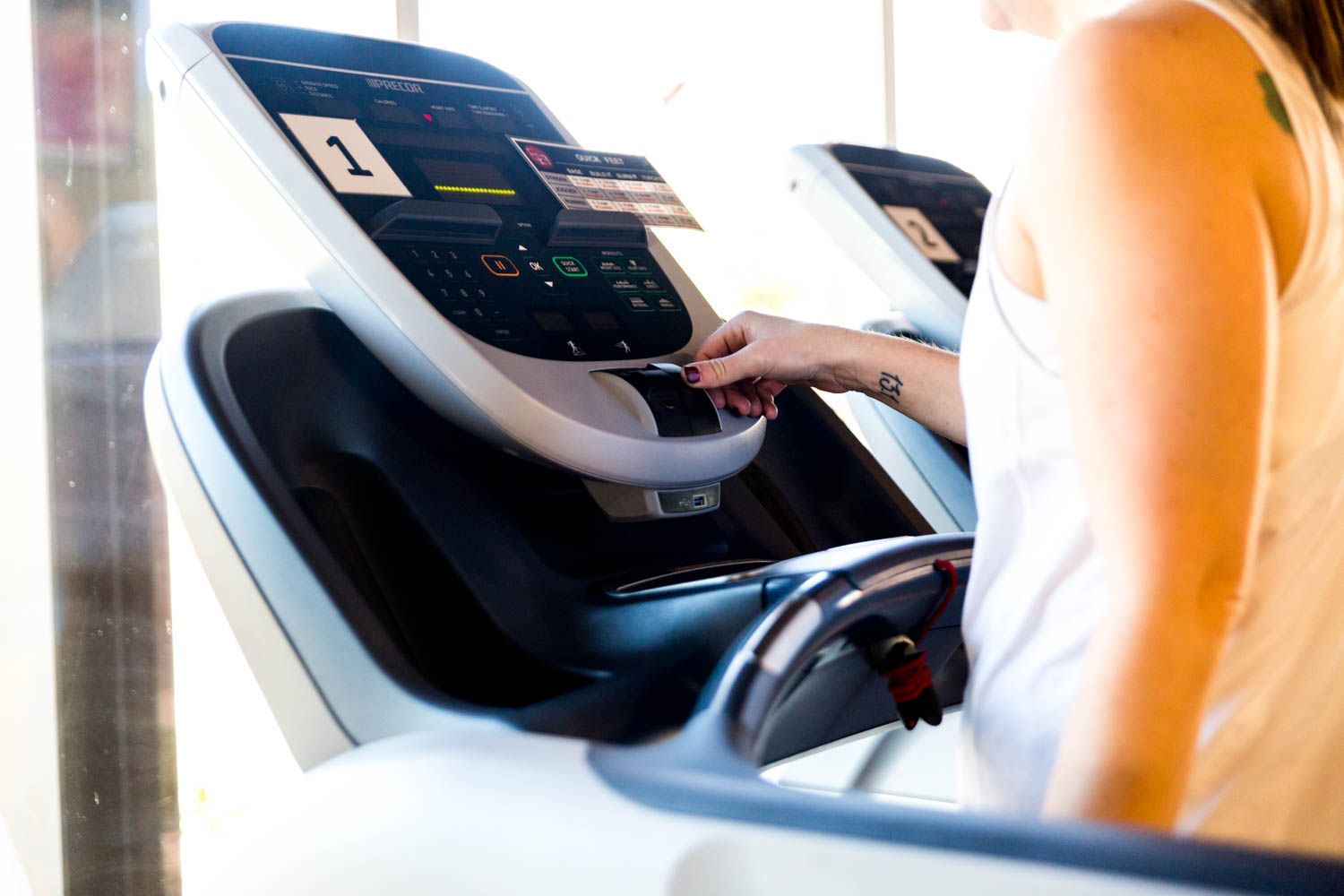 Person using a treadmill's touchscreen controls in a gym. White tank top, arm with tattoo.