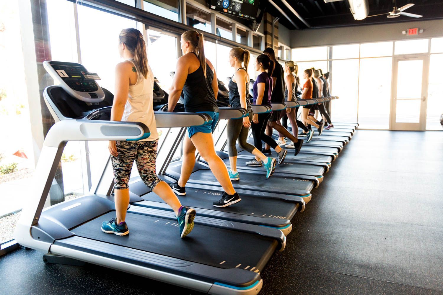 People running on treadmills in a brightly lit gym next to a large window.