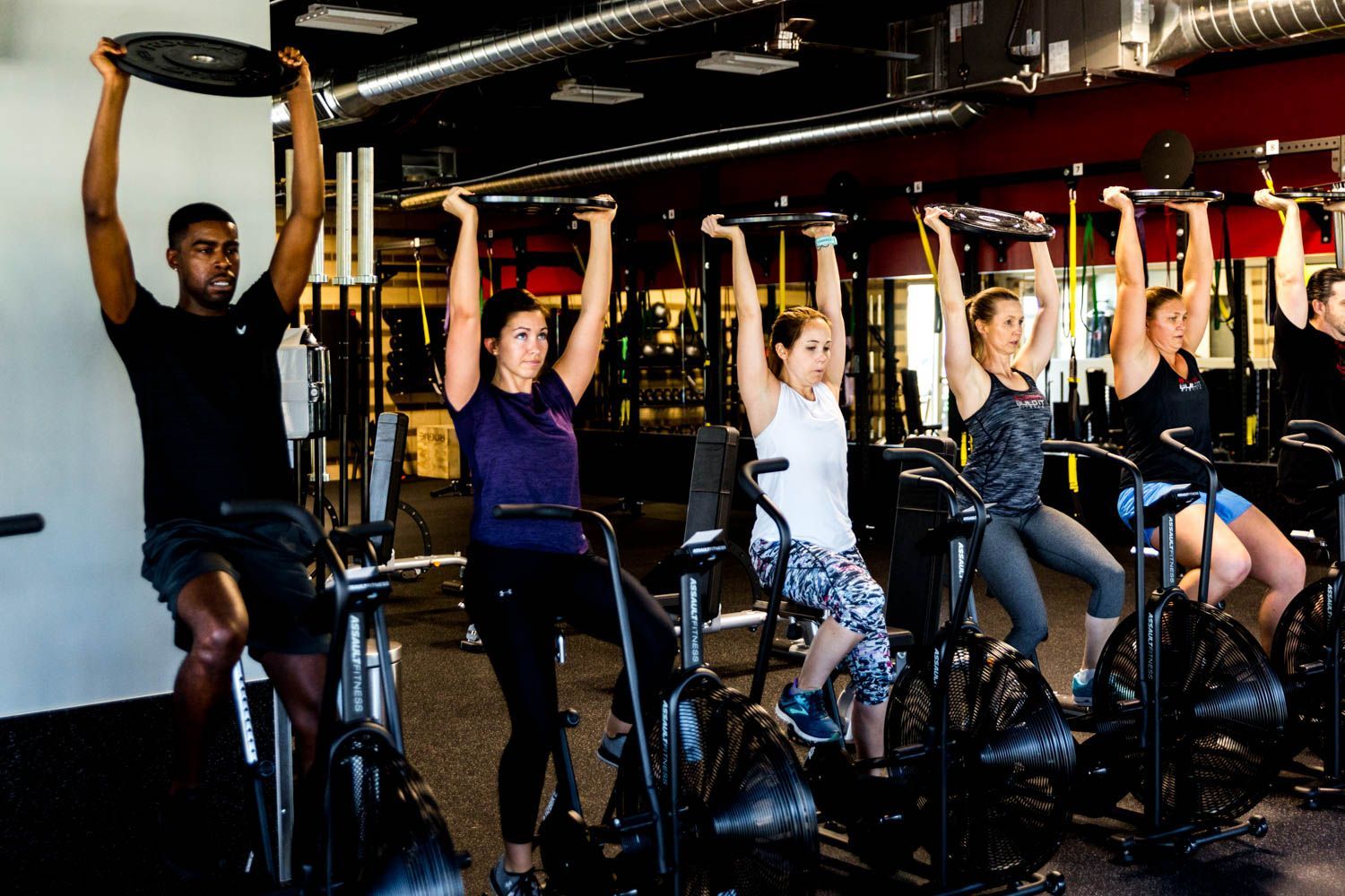 People working out on stationary bikes, holding weights above their heads. Gym setting.
