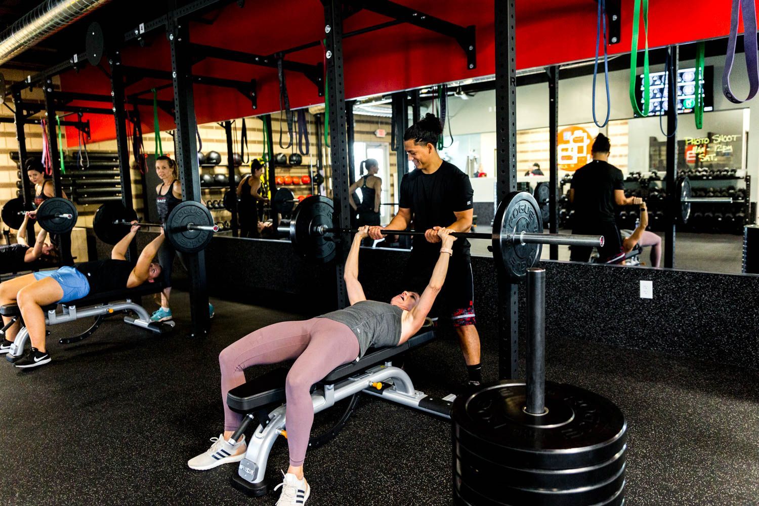 People lifting weights in a gym. Woman bench presses with spotter; other people exercise nearby. Dark interior.