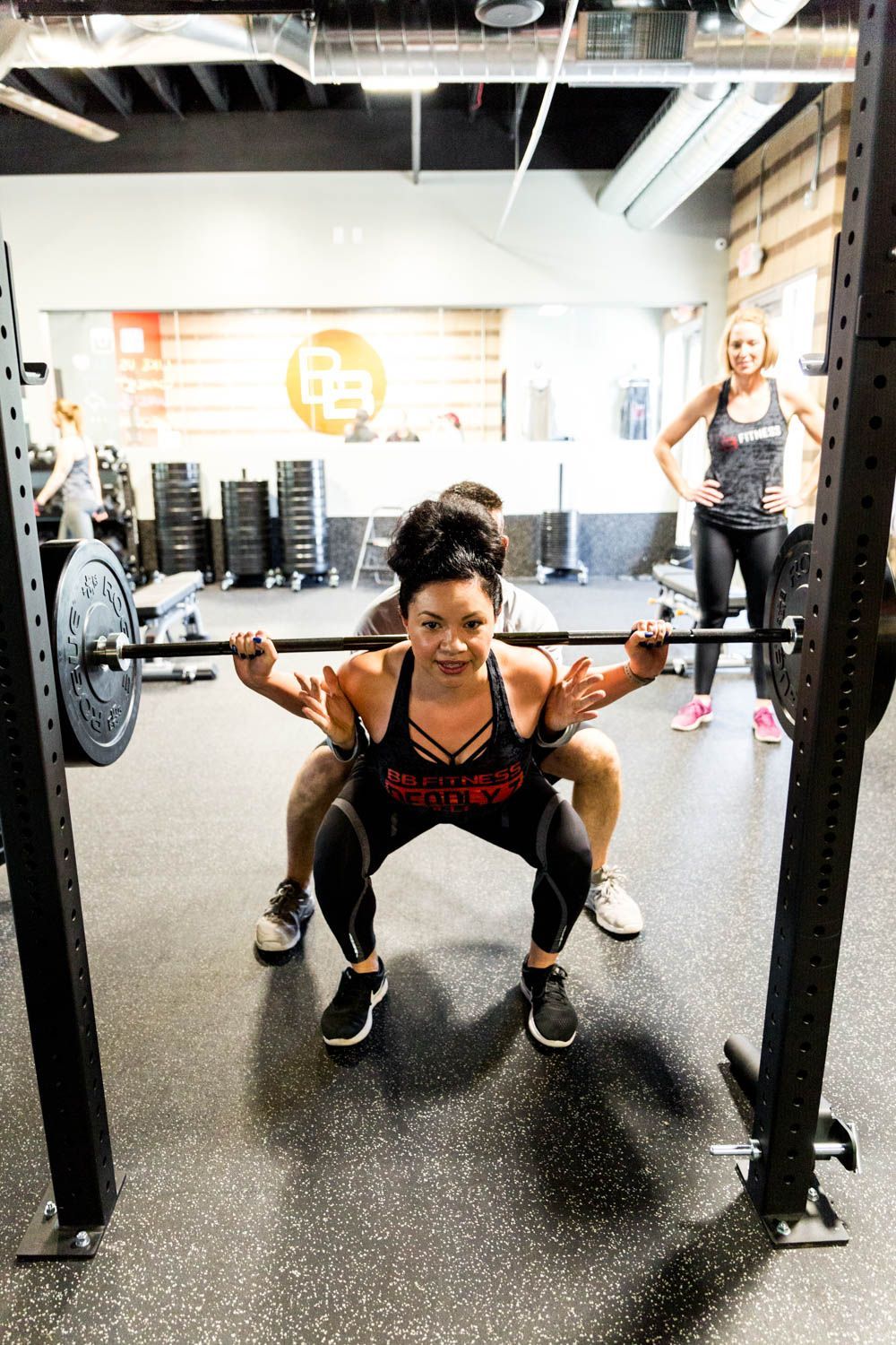 Woman squats with barbell, assisted by a person. Another person watches in a gym setting.