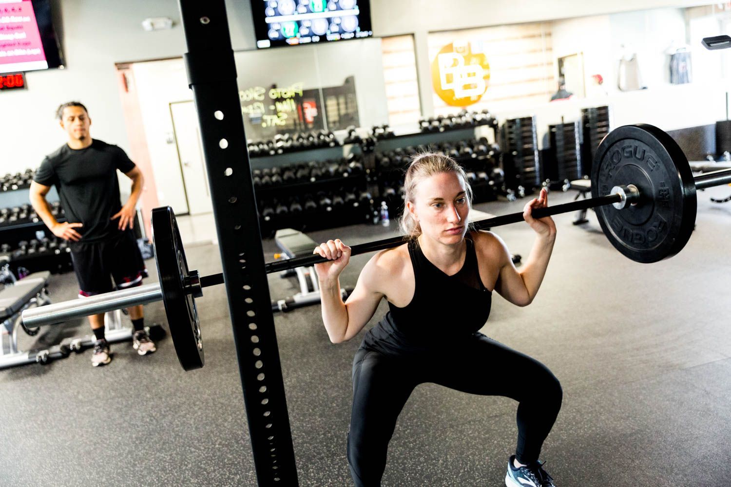 Woman squatting with barbell in a gym, man watching in background.