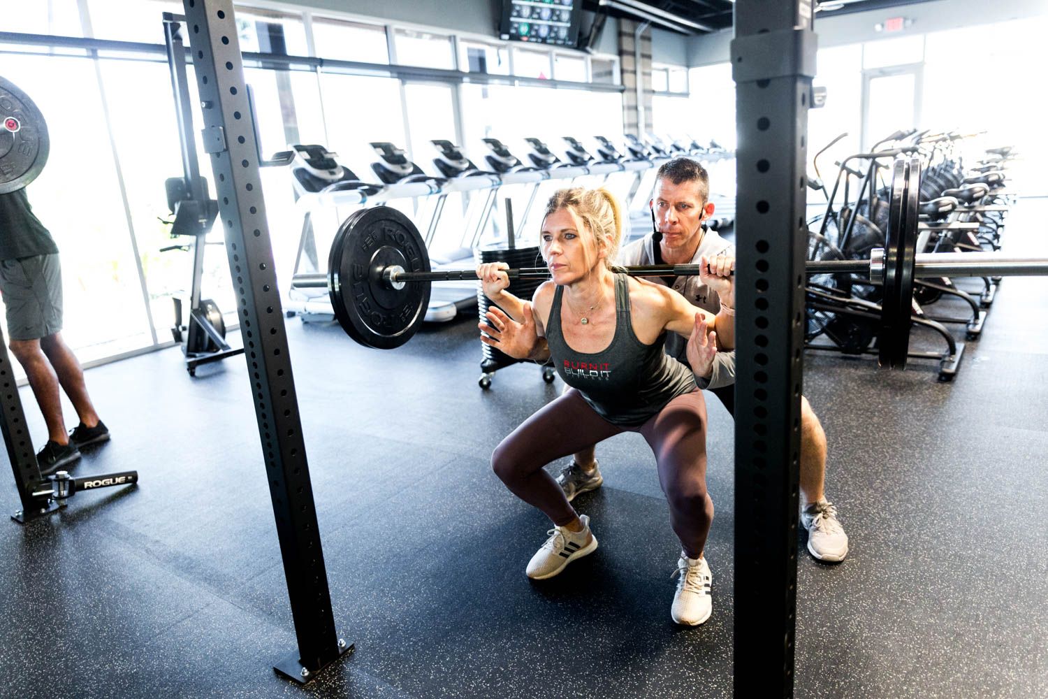 Woman squatting with barbell, being spotted by trainer in gym.