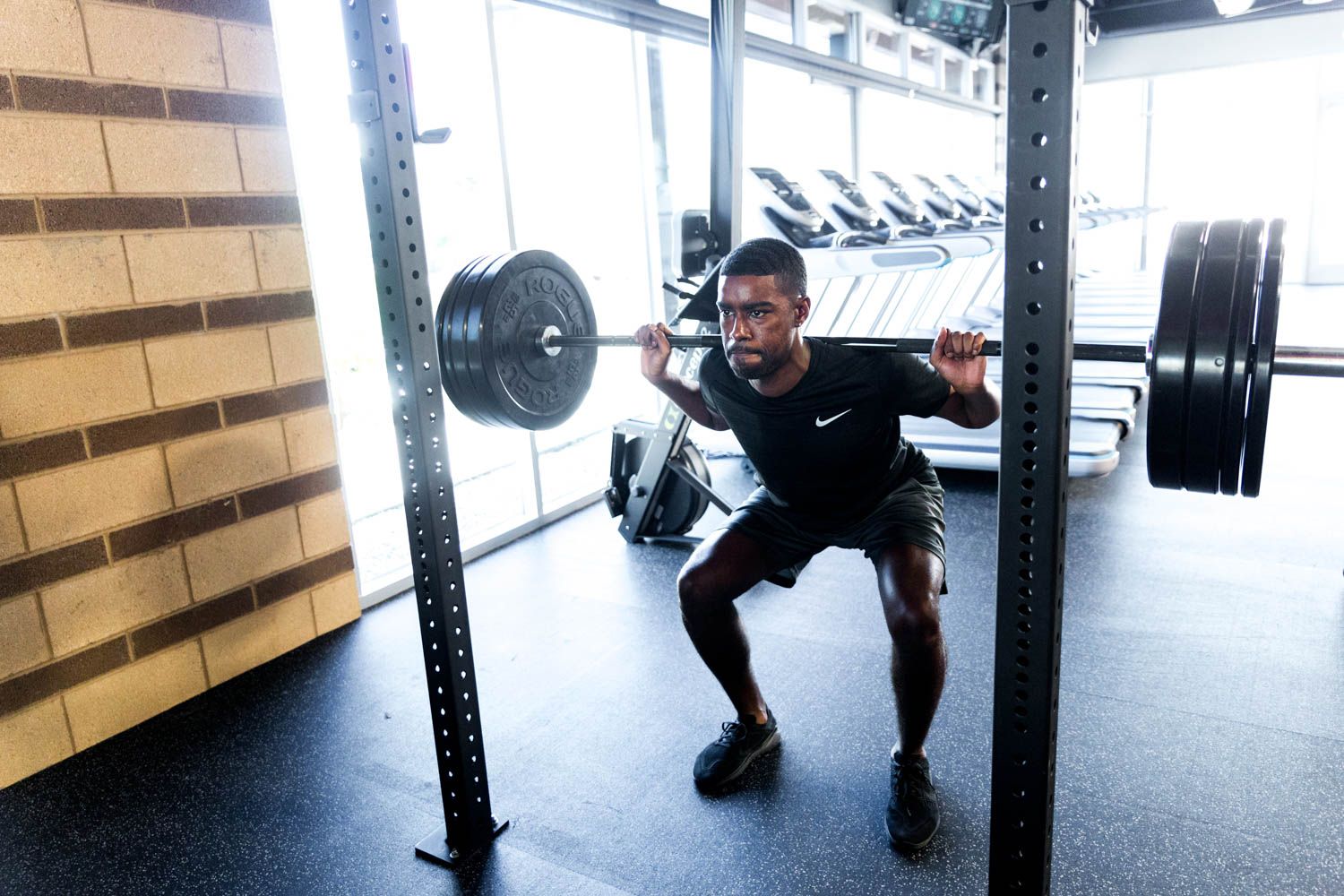 Man performing barbell squat in a gym. Black t-shirt, shorts, dark shoes. Facing forward, eyes focused.