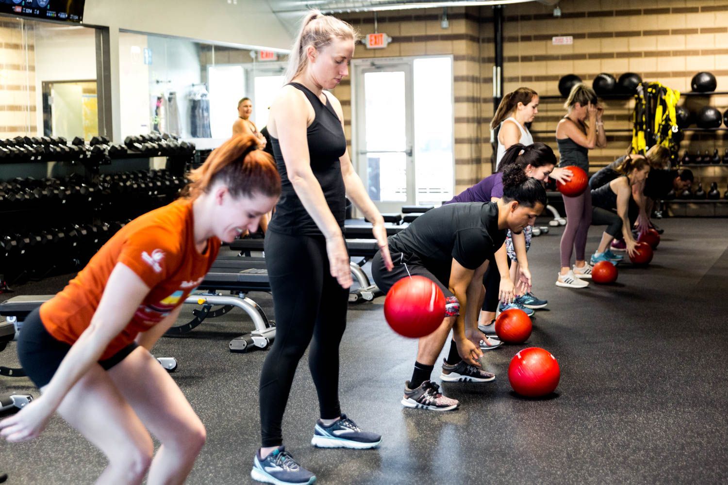 A fitness instructor guides a group doing medicine ball squats in a gym.