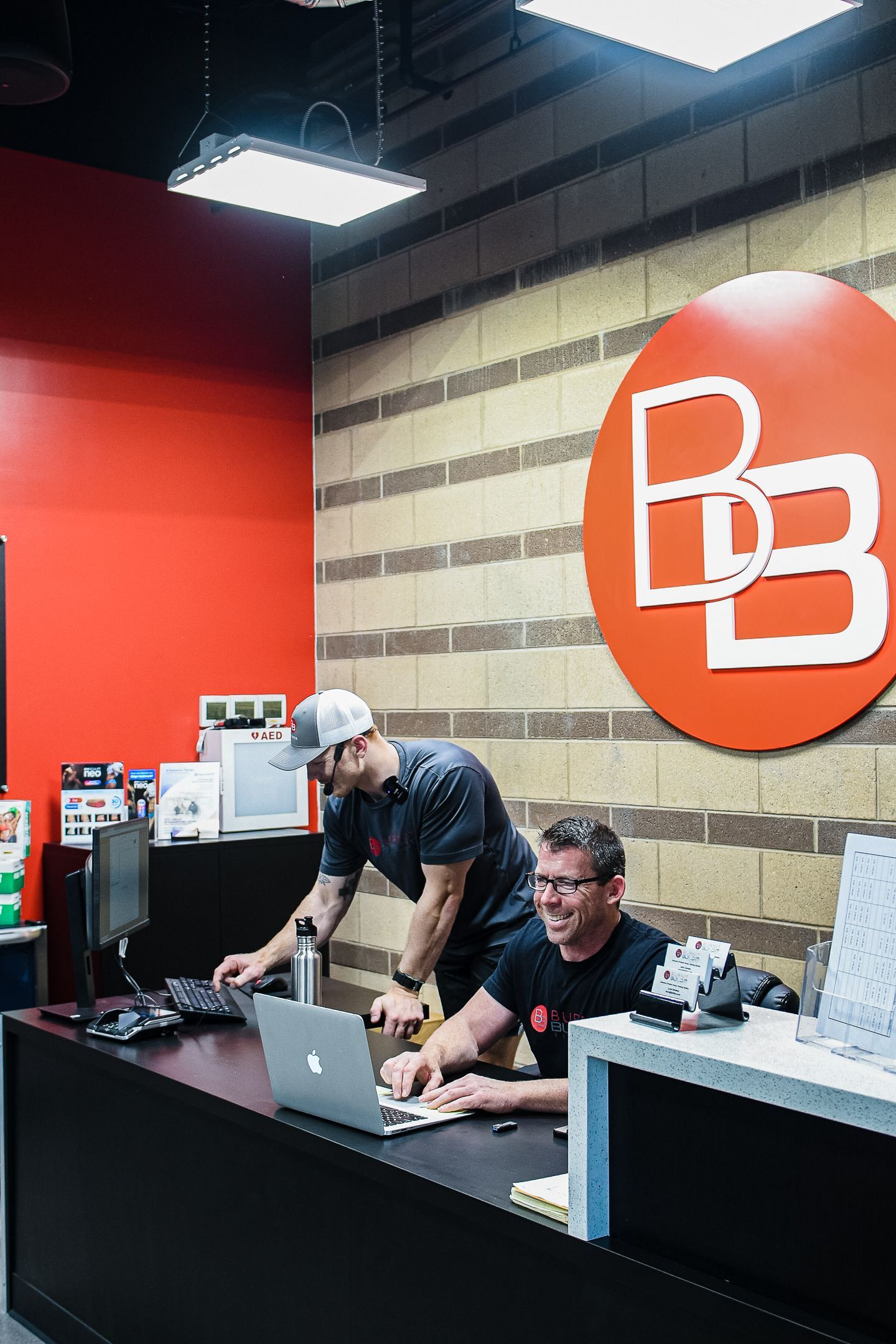 Two men at a reception desk with a laptop and a logo. One is working, another behind him. Red and brick wall background.