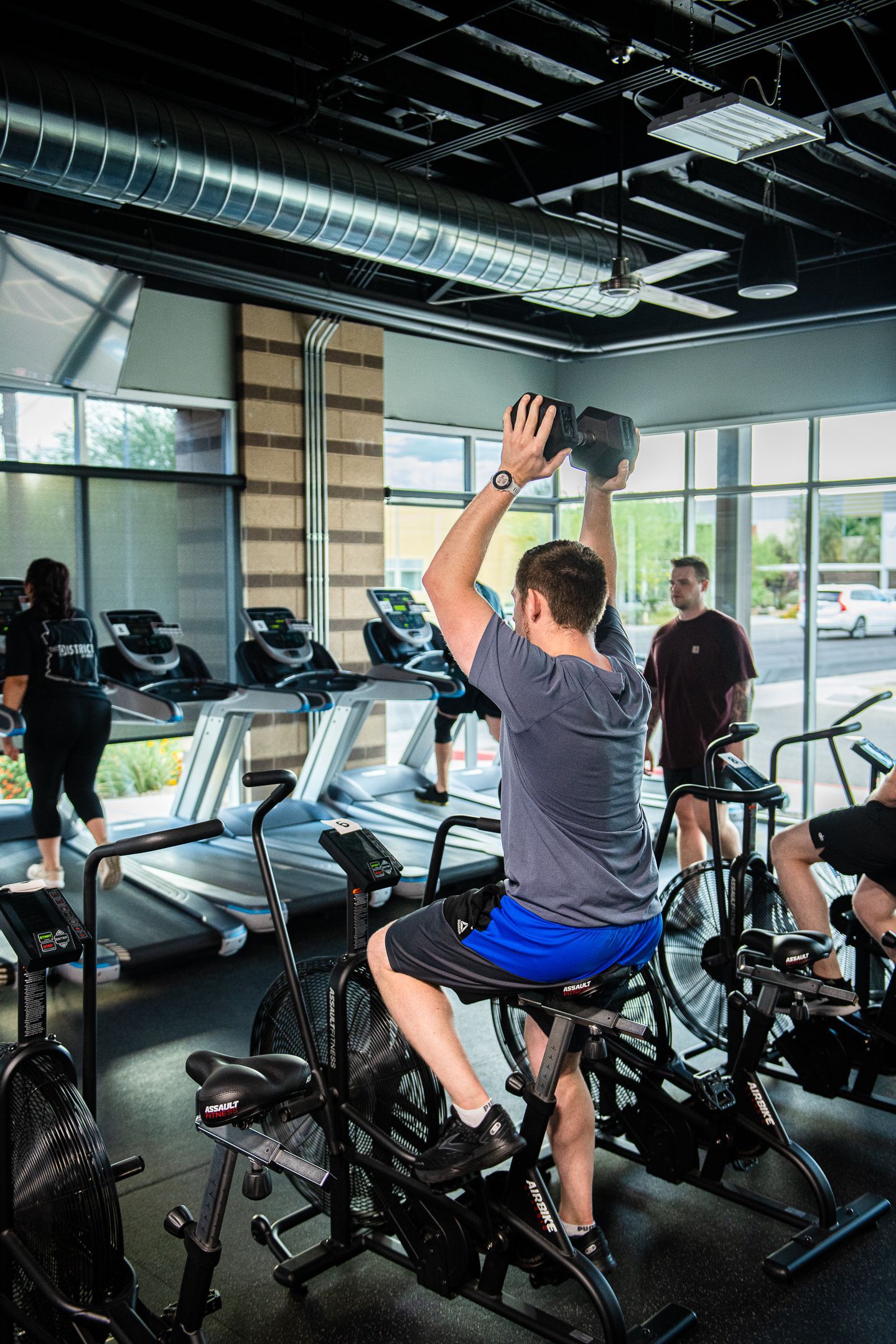 Man working out on a stationary bike, holding dumbbells over his head, at a gym with other people using exercise equipment.