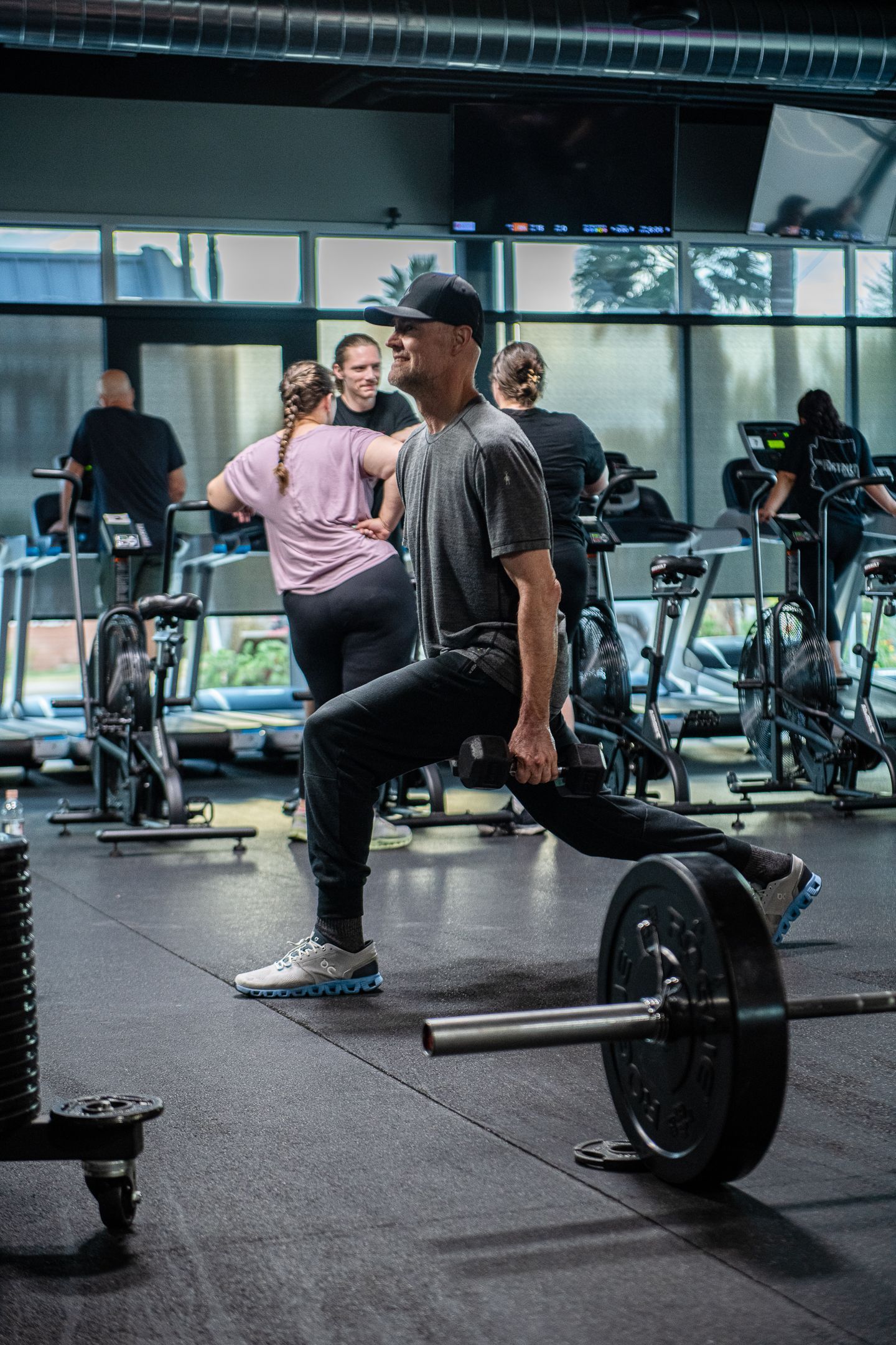 Man lunges with barbell in a gym. Others use cardio machines in background.