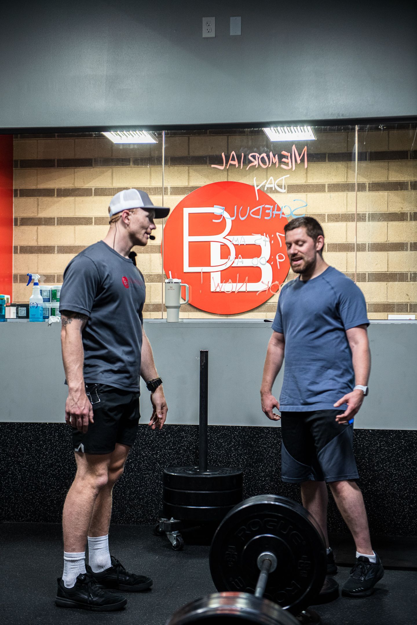 Two men in a gym. One wearing hat and shorts stands near a barbell, the other in a t-shirt looks on.