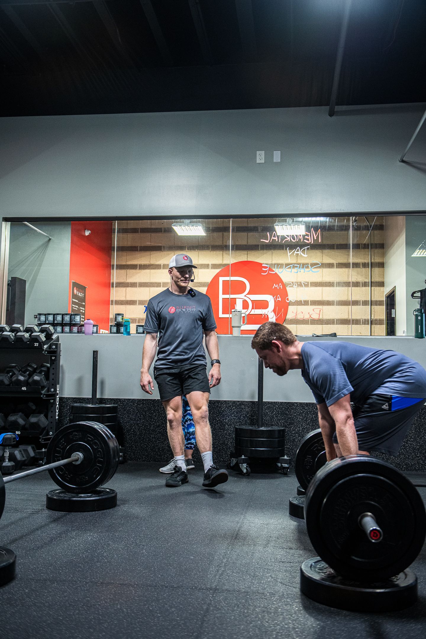 Man lifting barbell, supervised by a coach in a gym with weights and a logo on the wall.