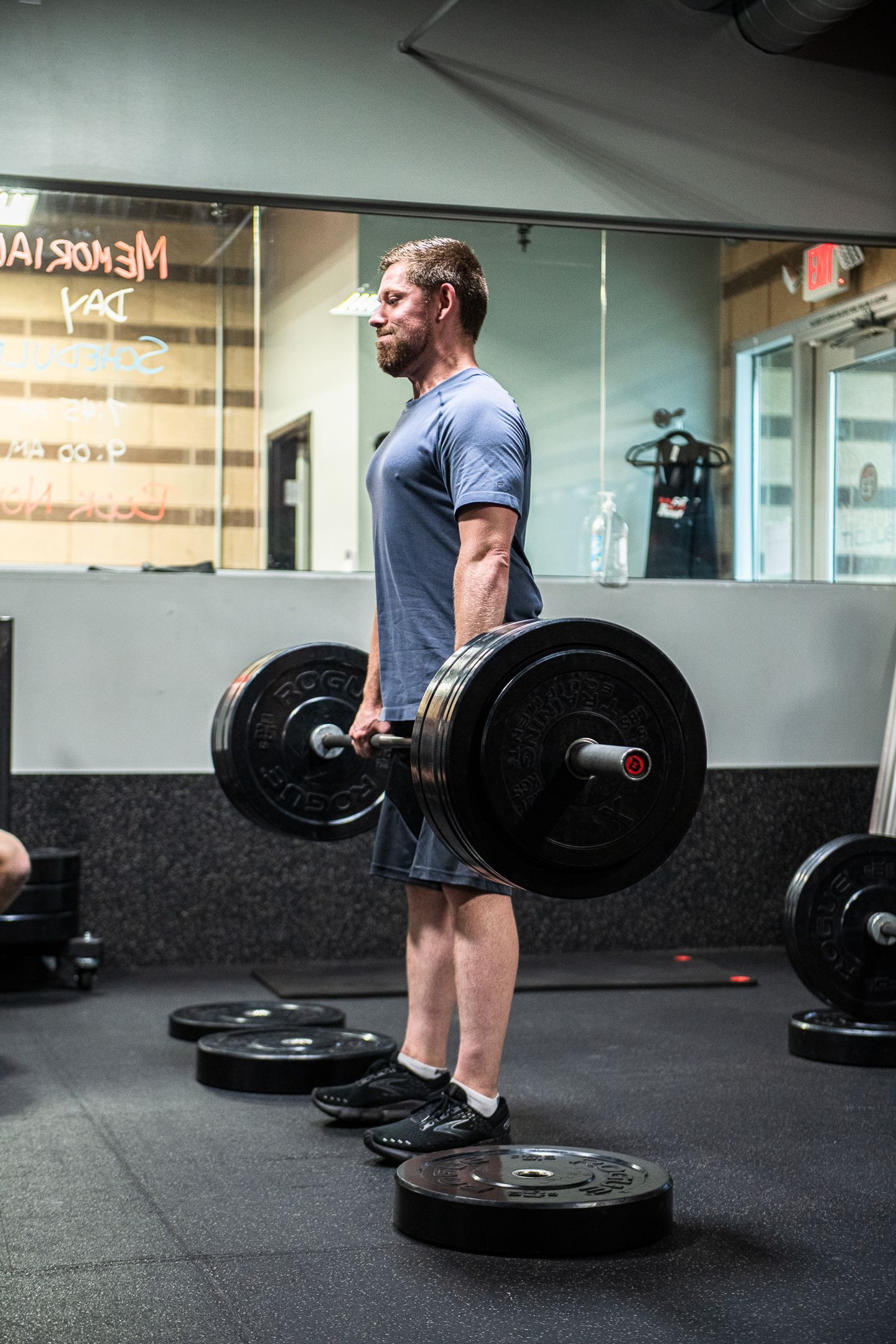 Man lifting a barbell in a gym, standing on plates. Dark clothing, serious expression.