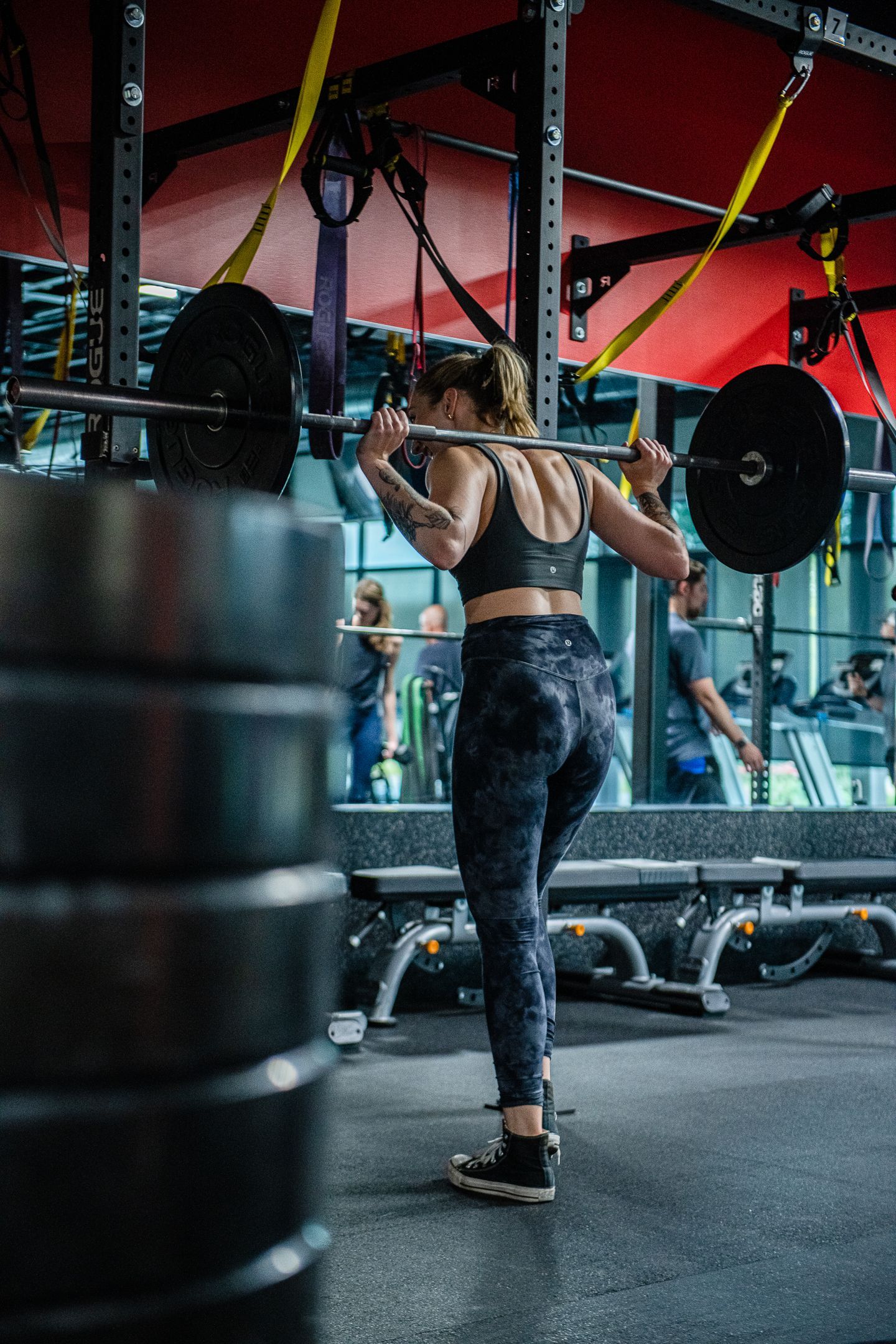 Woman performing a barbell squat in a gym, facing a mirror. She wears athletic wear, weights are stacked.