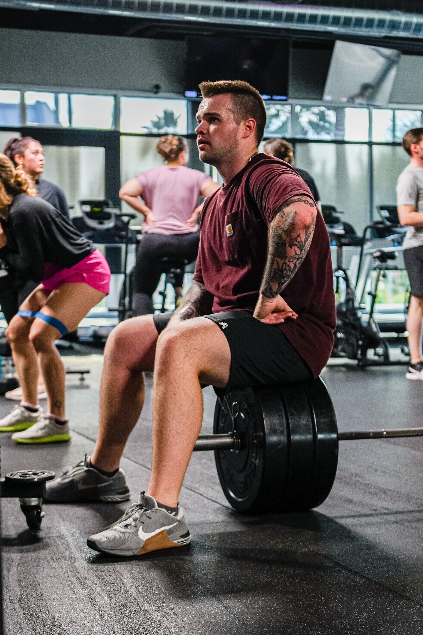 Man sitting on weight, barbell across legs, inside gym. Others exercise in background.