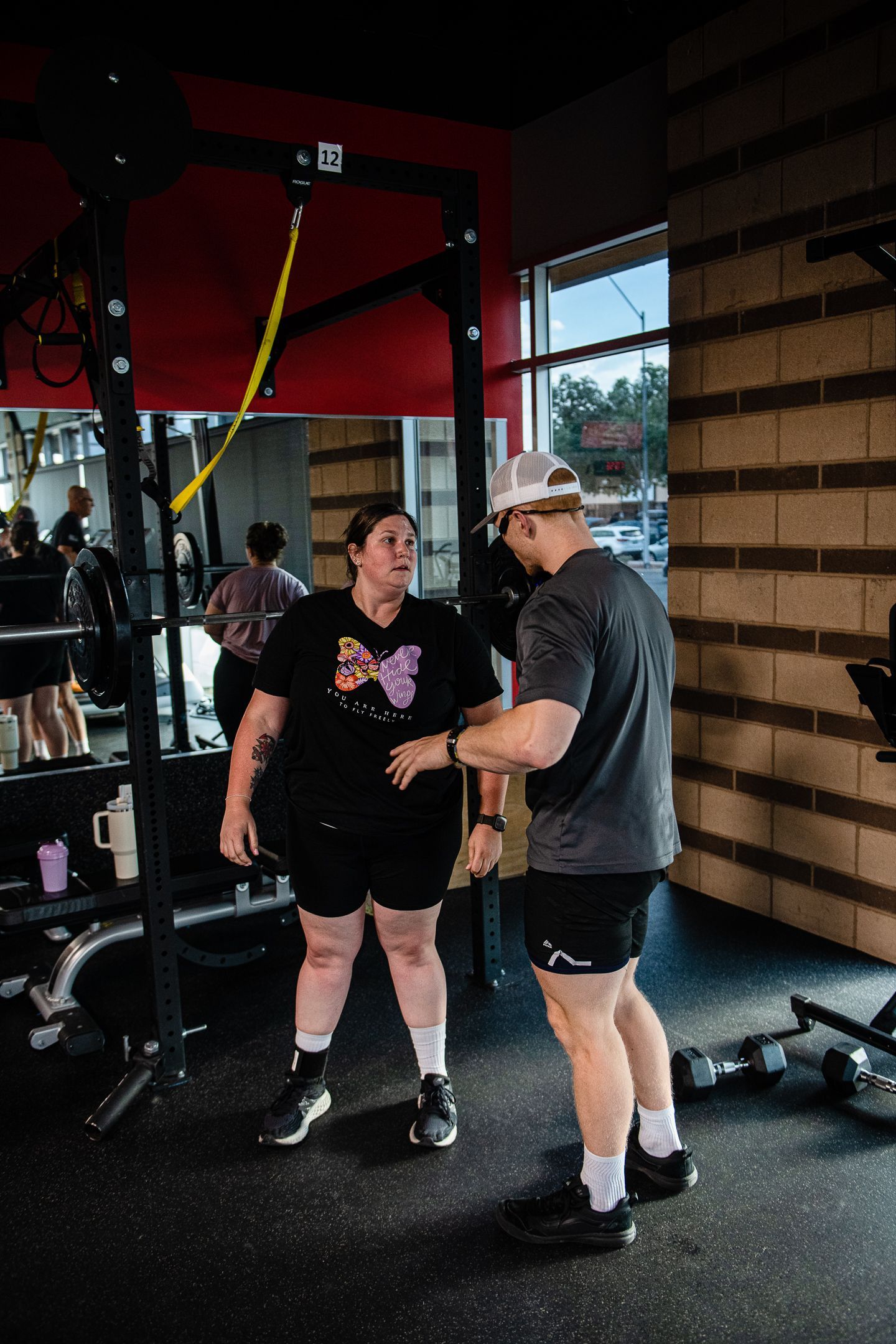 A person in black shorts and a t-shirt talks with a person who wears a hat in a gym.