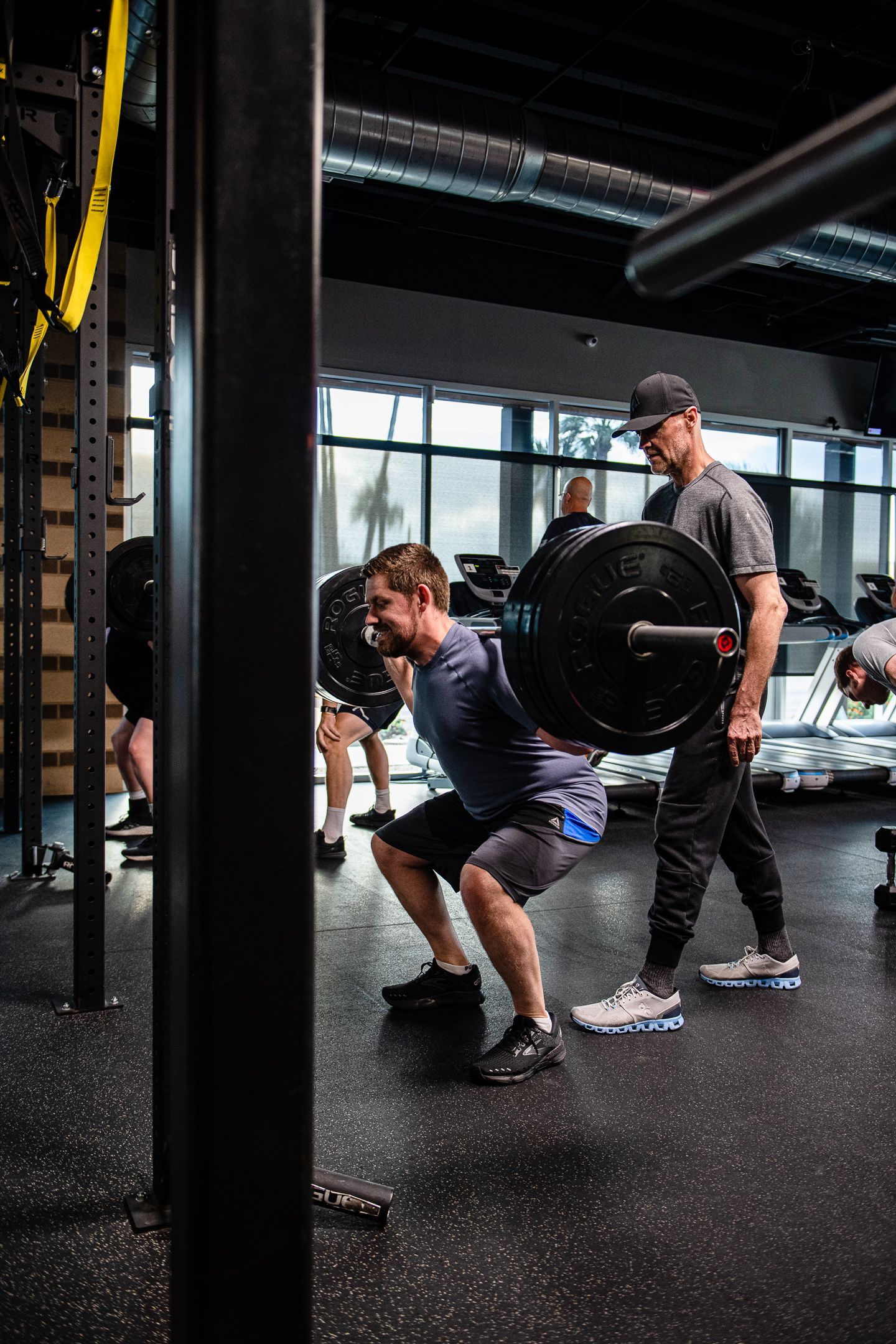 Man squatting with barbell, instructor spotting in modern gym.