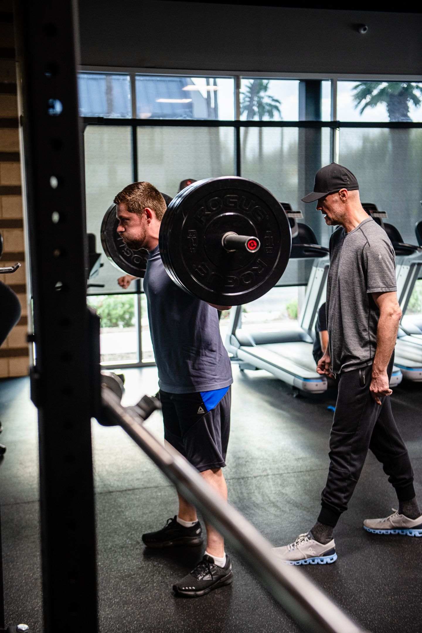 Man squatting with barbell, spotter observing in gym, with weightlifting rack and treadmills in background.