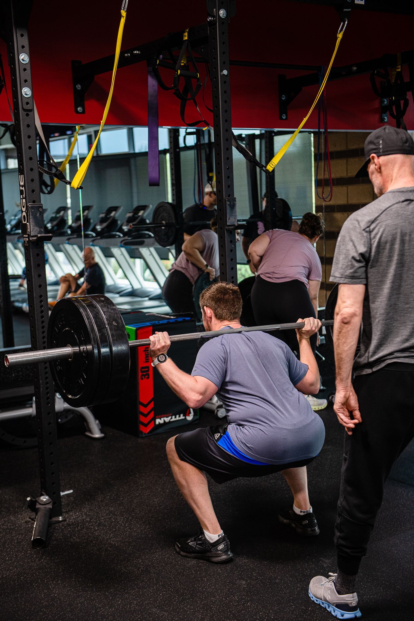 Man squats with barbell, guided by a coach in a gym. Other people exercise in the background.