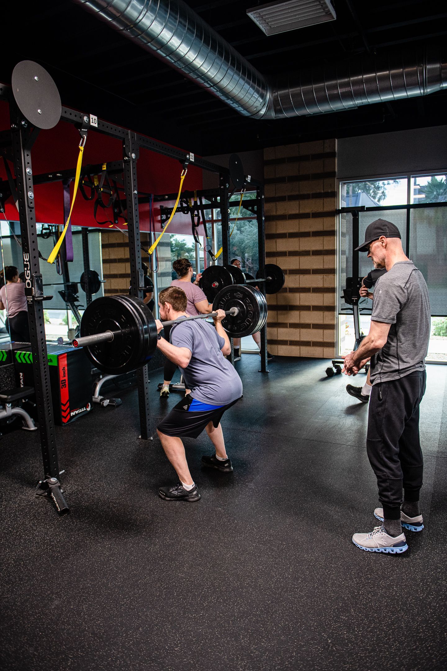 Man performing a barbell squat in a gym, with a trainer observing.