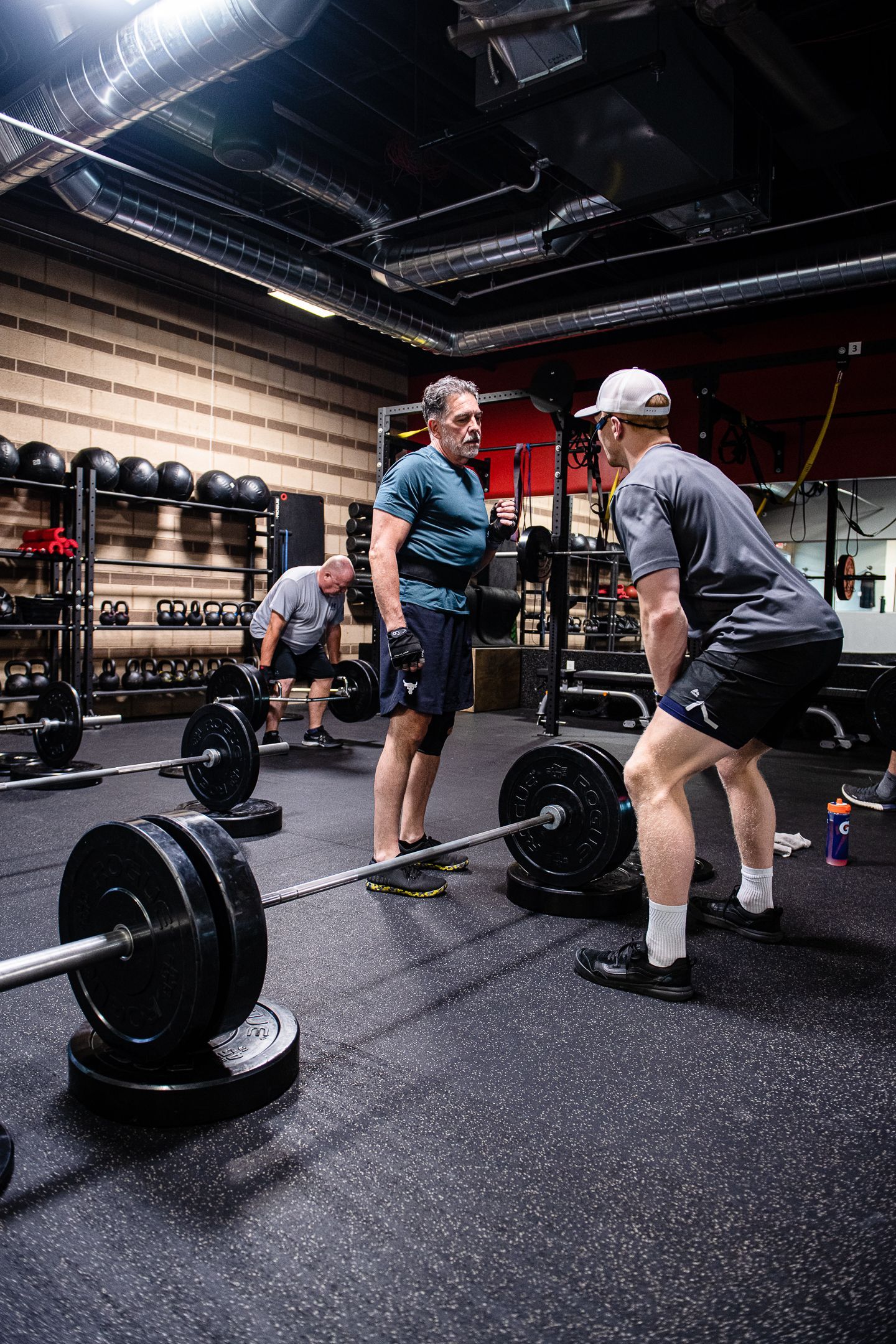 Two men in gym setting, one lifting barbell while another spots. Black weights, rubber floor.