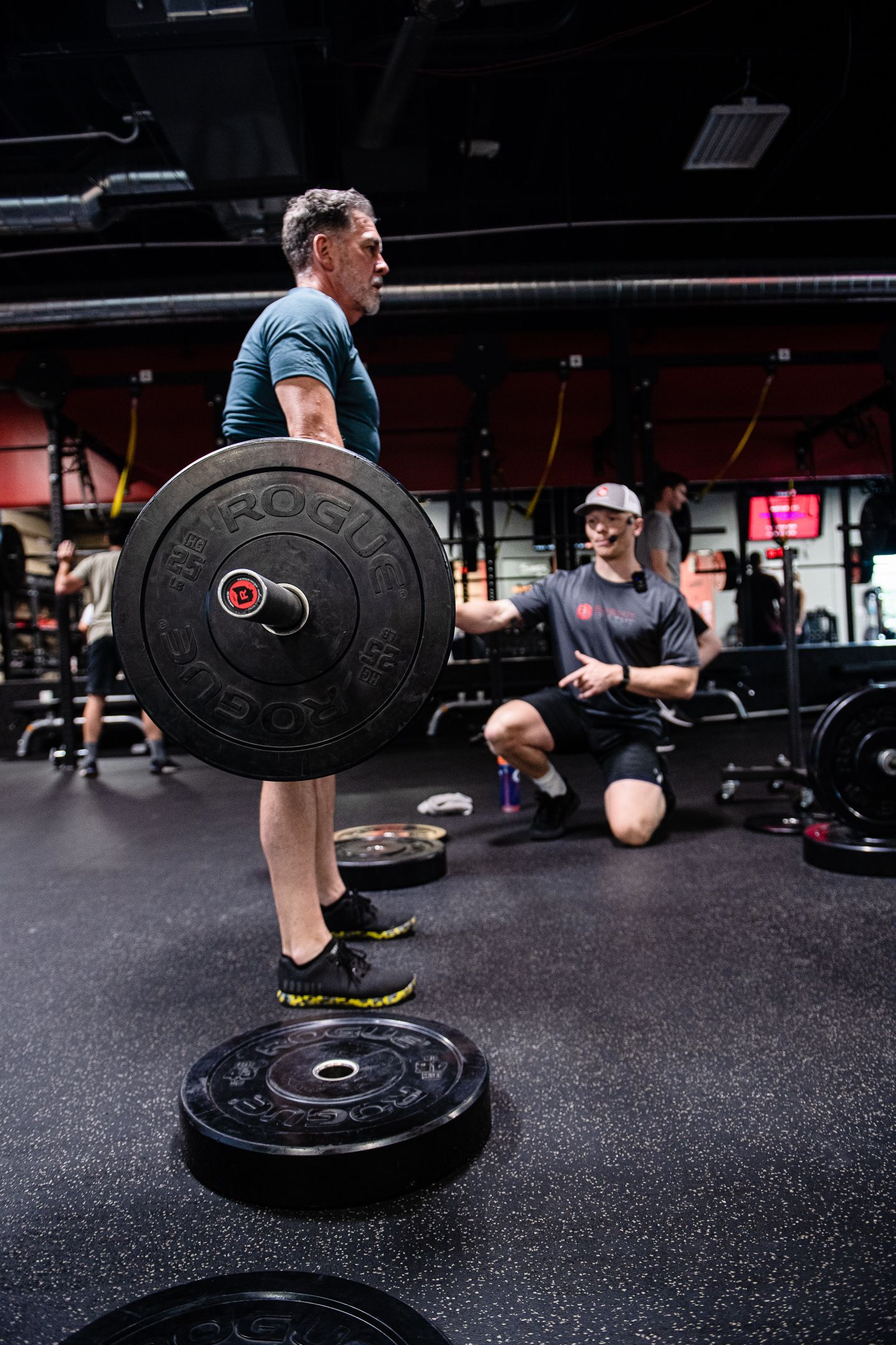 Man lifting barbell in gym, coach watching. Black weights on floor, dark setting.