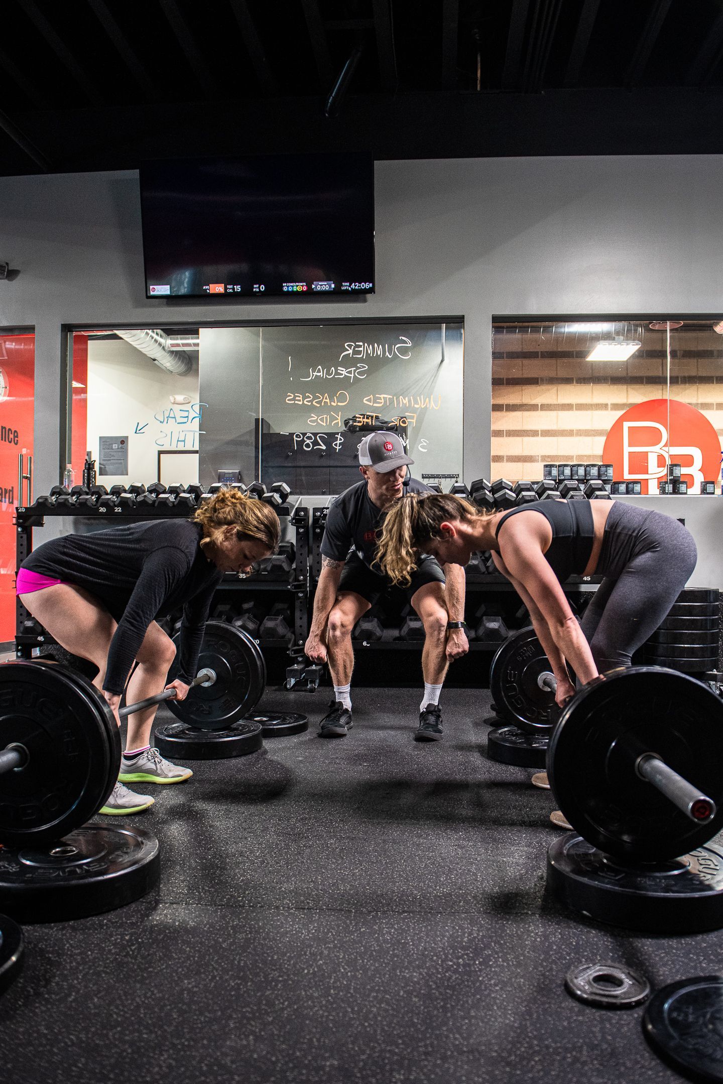 Three people lifting barbells in a gym. Black flooring, weights, and a TV are visible.