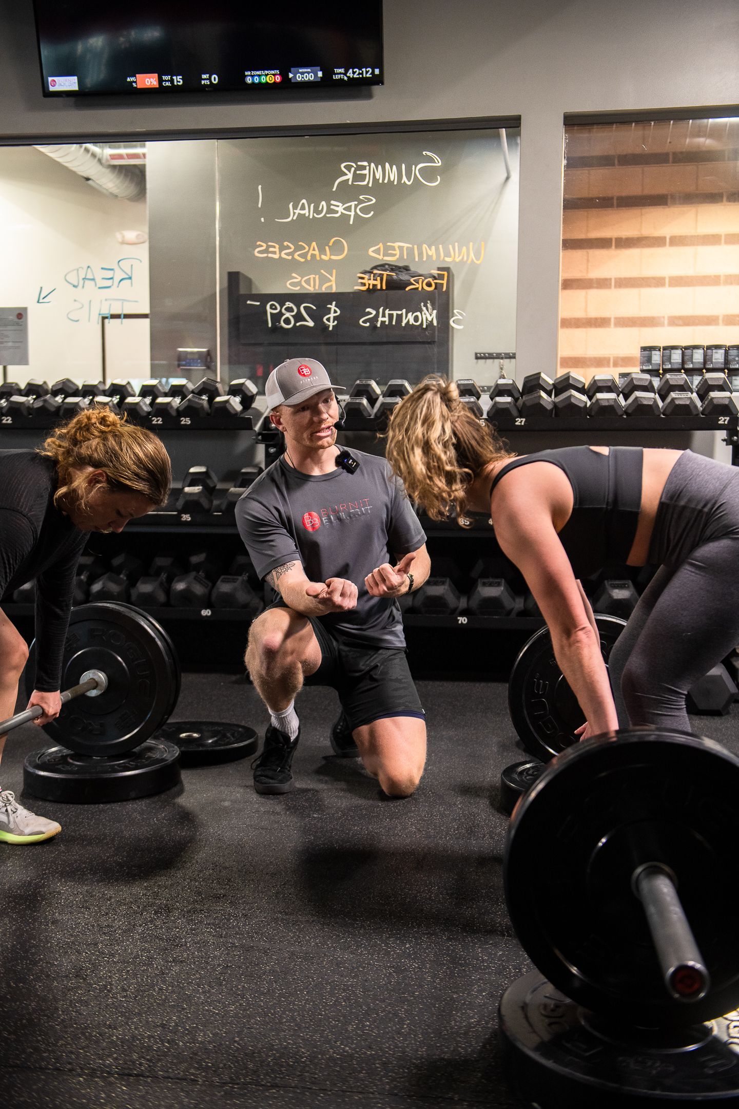 Trainer coaching two people lifting weights in a gym. Black barbell plates, mirrored wall, and dark workout floor.