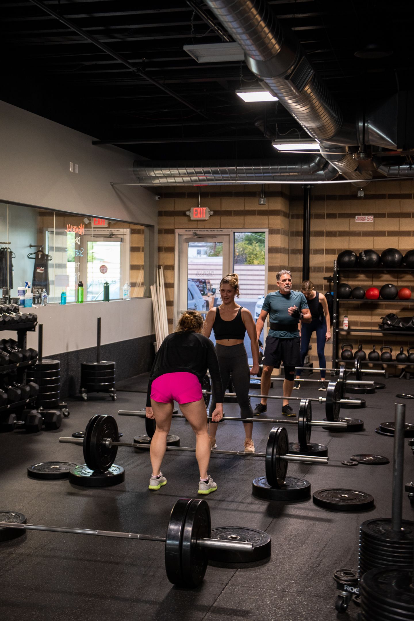 People working out in a gym, lifting barbells. Several others observe. Dark-toned interior with weight equipment.