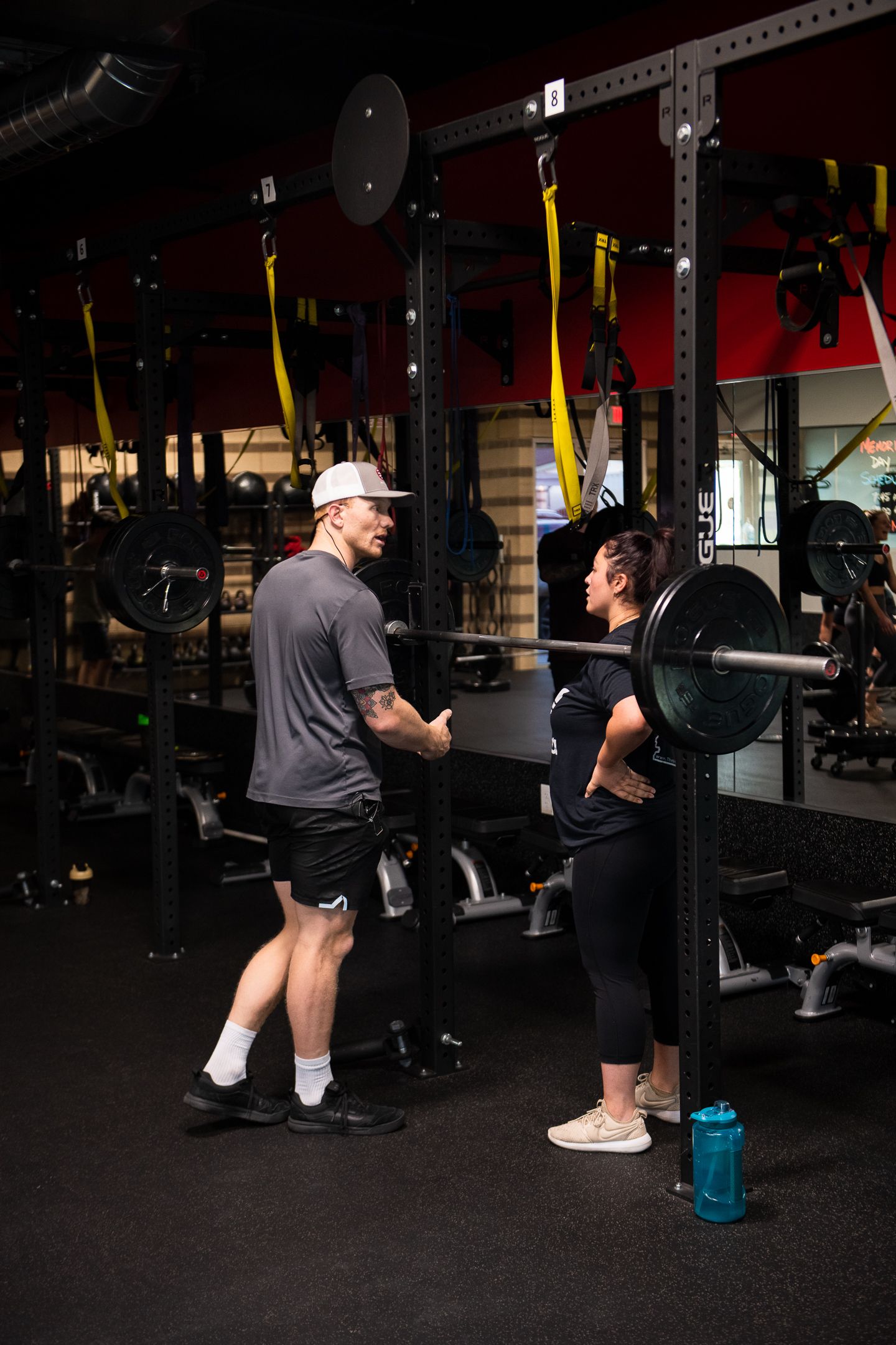 A person squats with a barbell while a trainer stands by in a gym.