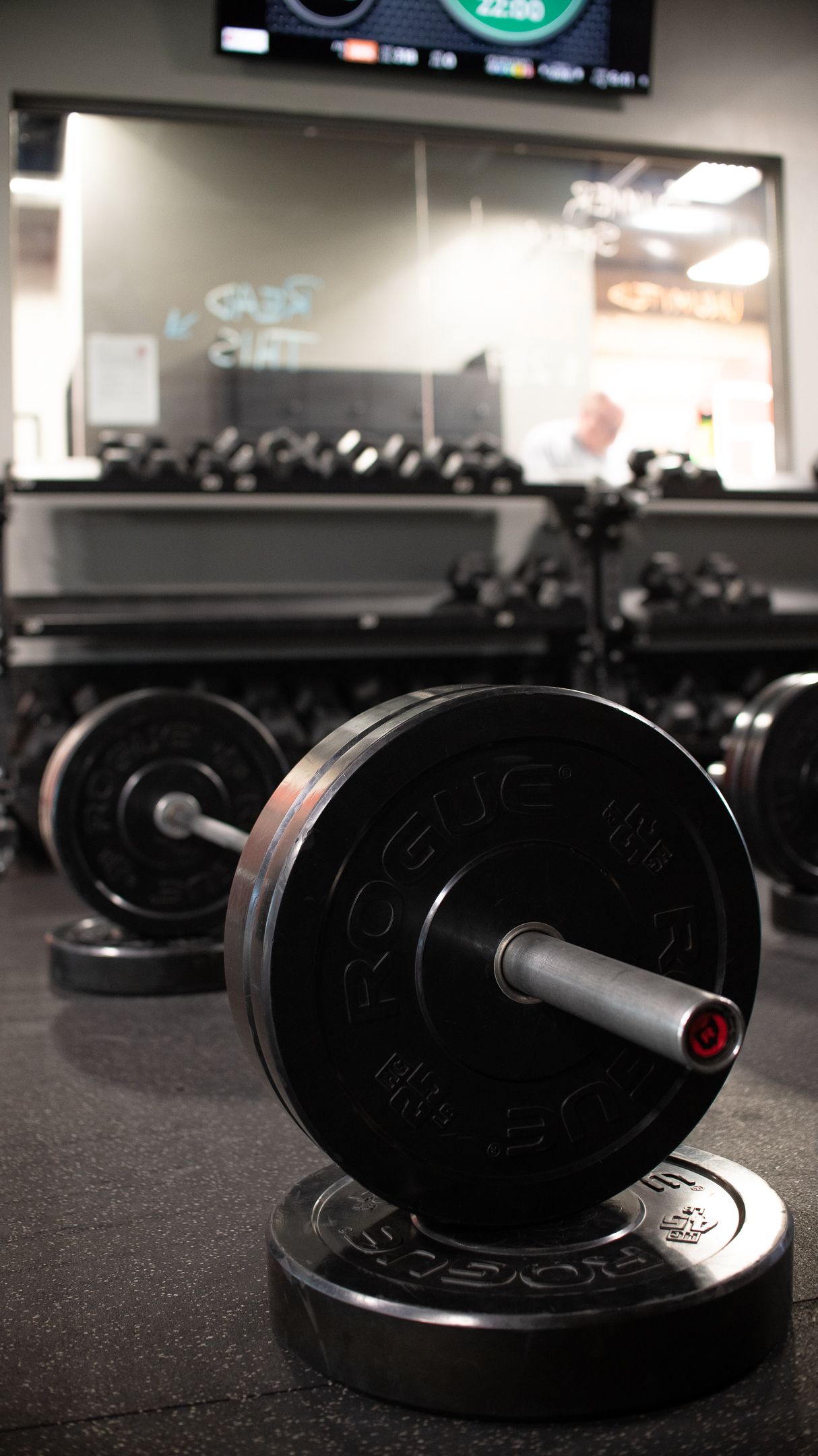 Barbell loaded with black weight plates on a gym floor, with dumbbells and a clock in the background.