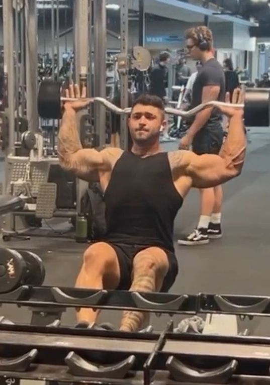 Man seated, lifting barbell overhead at a gym. He wears black and has arm tattoos. A person observes nearby.