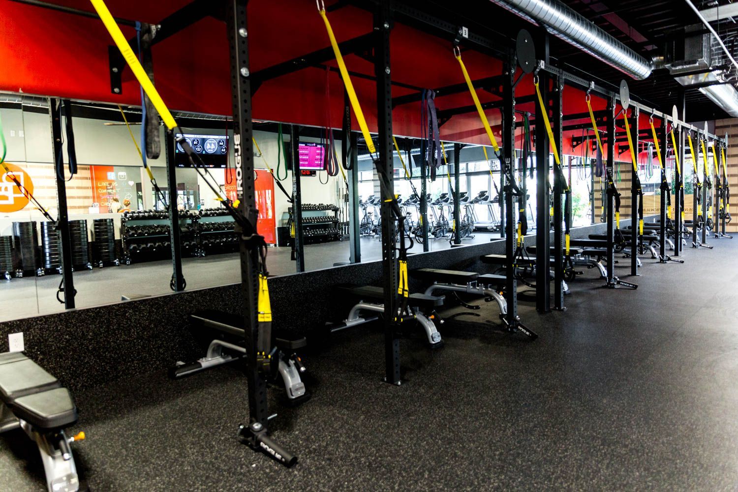 Gym interior with TRX suspension trainers, benches, dumbbells, and mirrors. Black flooring and equipment with red and yellow accents.