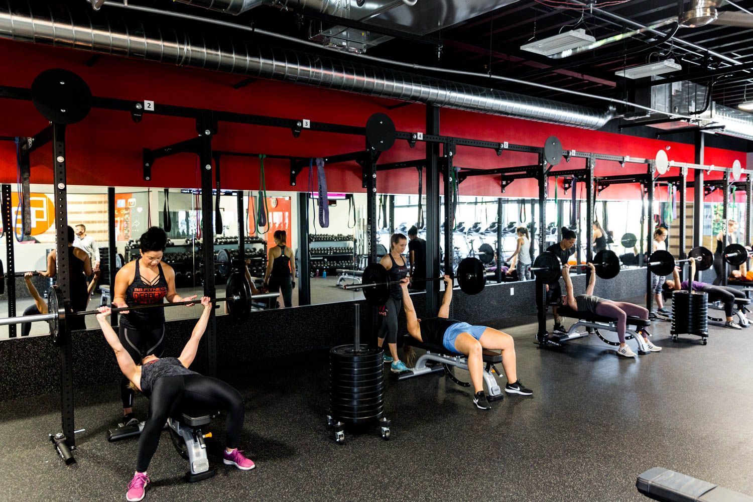 People lifting weights at a gym. Reflective mirrors, black equipment, red walls.