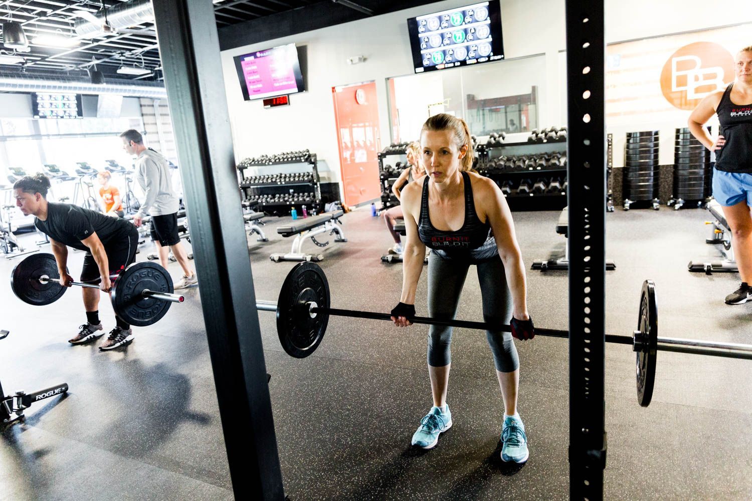 People lifting weights in a gym. Woman in black activewear with barbell, others in background.